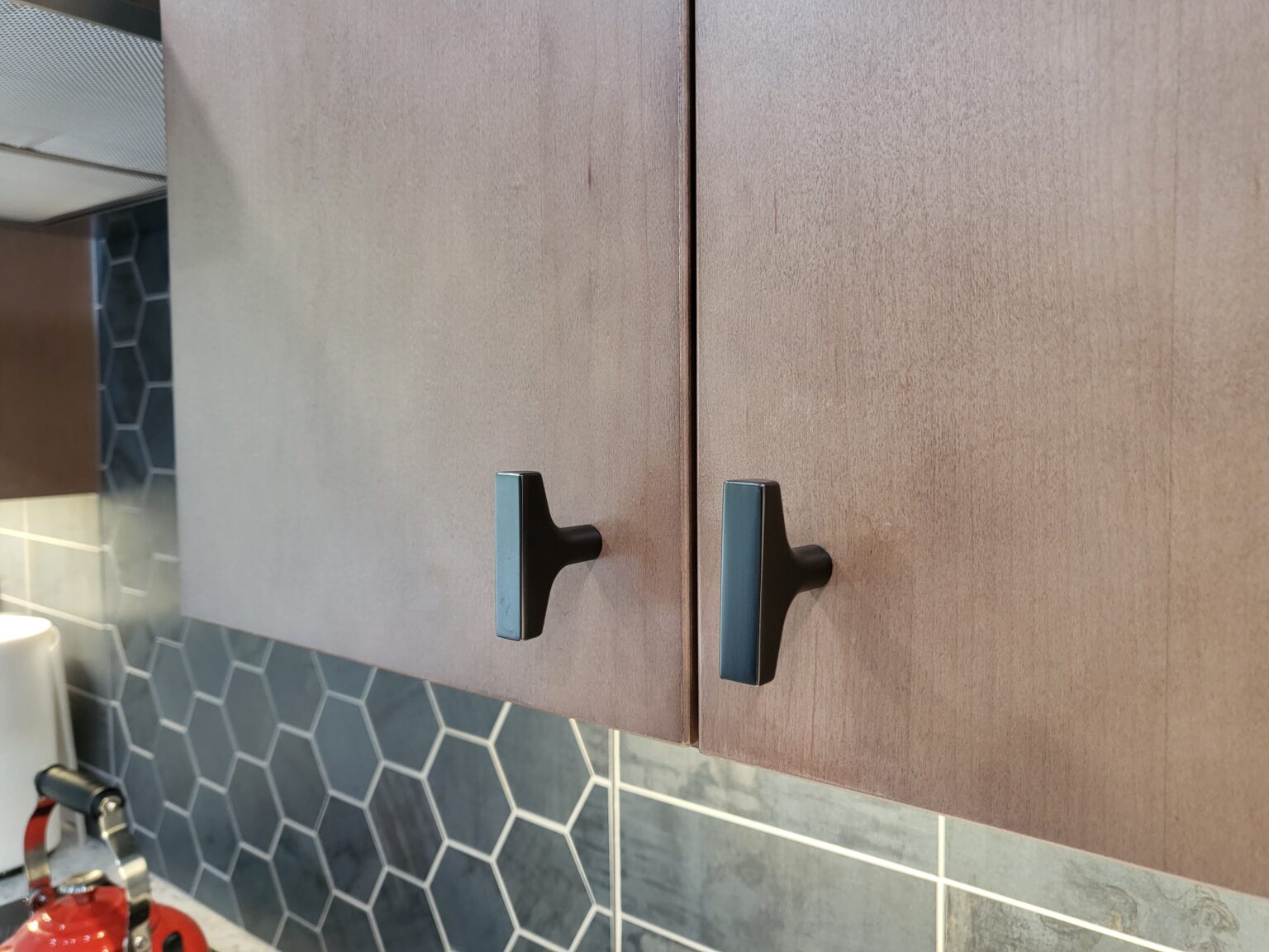 Two wooden kitchen cabinets with black handles are mounted above a backsplash featuring dark hexagonal tiles. A red kettle is visible in the bottom left corner.