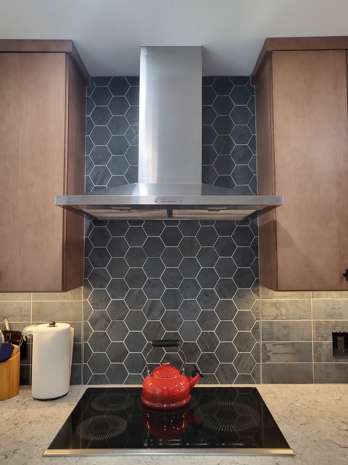 A red kettle sits on a black electric stovetop beneath a stainless steel range hood and hexagonal tile backsplash in a modern kitchen.