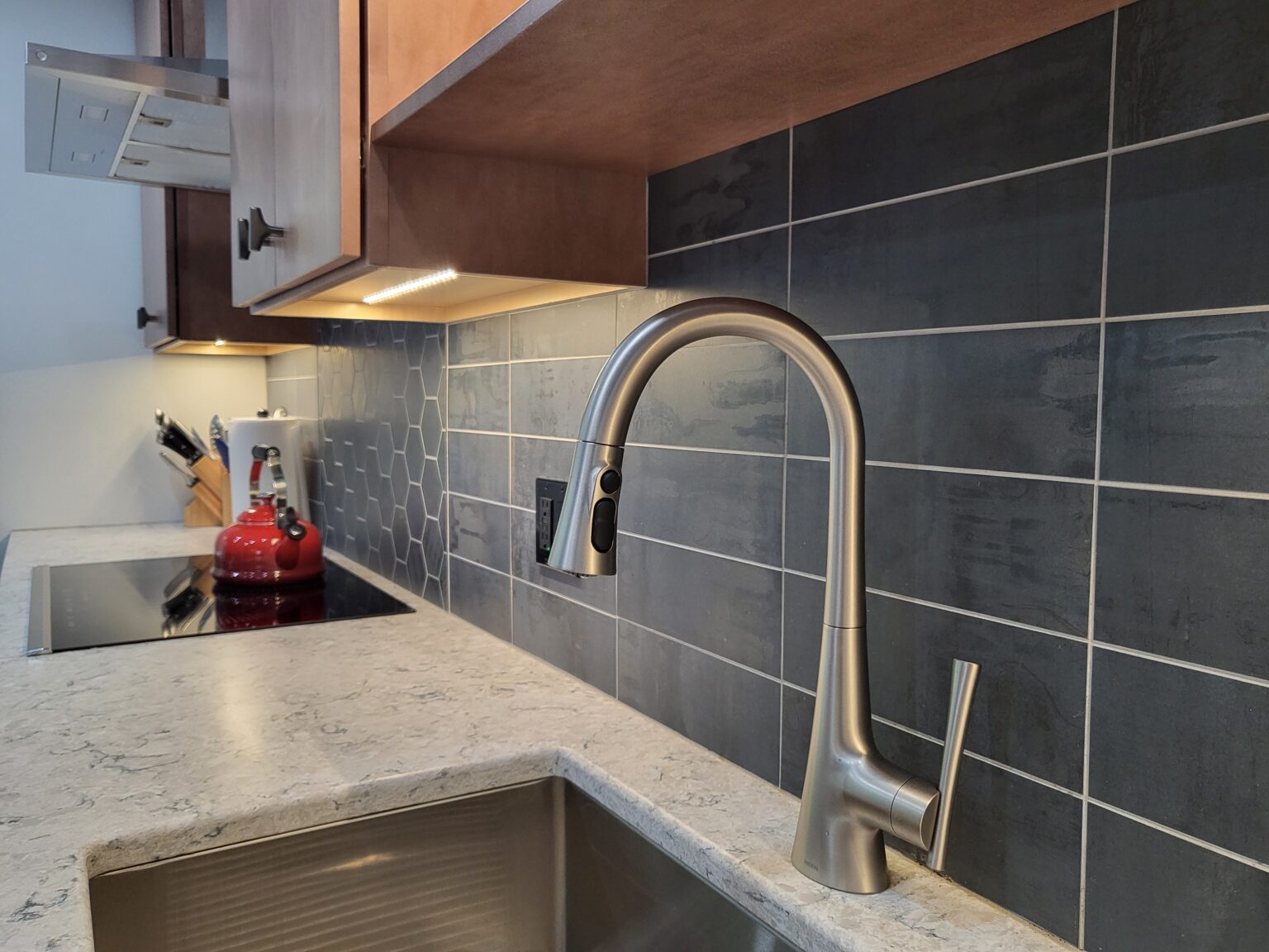 Modern kitchen with a stainless steel faucet, gray tiled backsplash, under-cabinet lighting, and a red kettle on the stove.