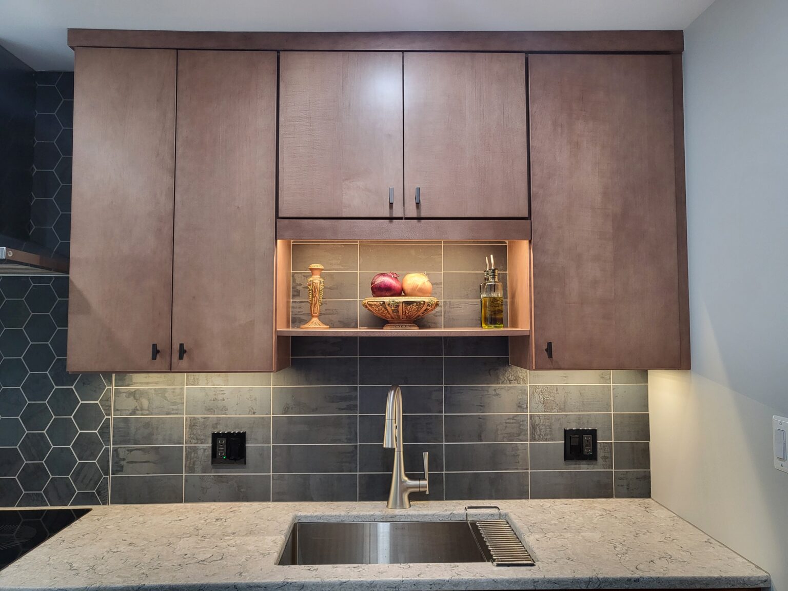 Modern kitchen with wood cabinets, a sink, and gray tile backsplash. Open shelf displays a bowl of onions and garlic, two glass bottles, and a candlestick.