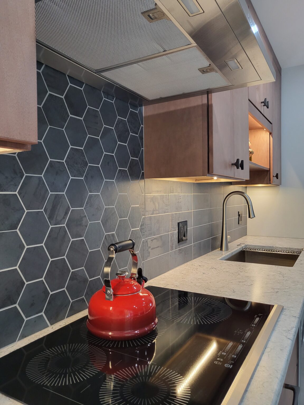 Modern kitchen with hexagonal tile backsplash, electric stovetop with a red kettle, light wood cabinets, undermount sink, and black faucet.