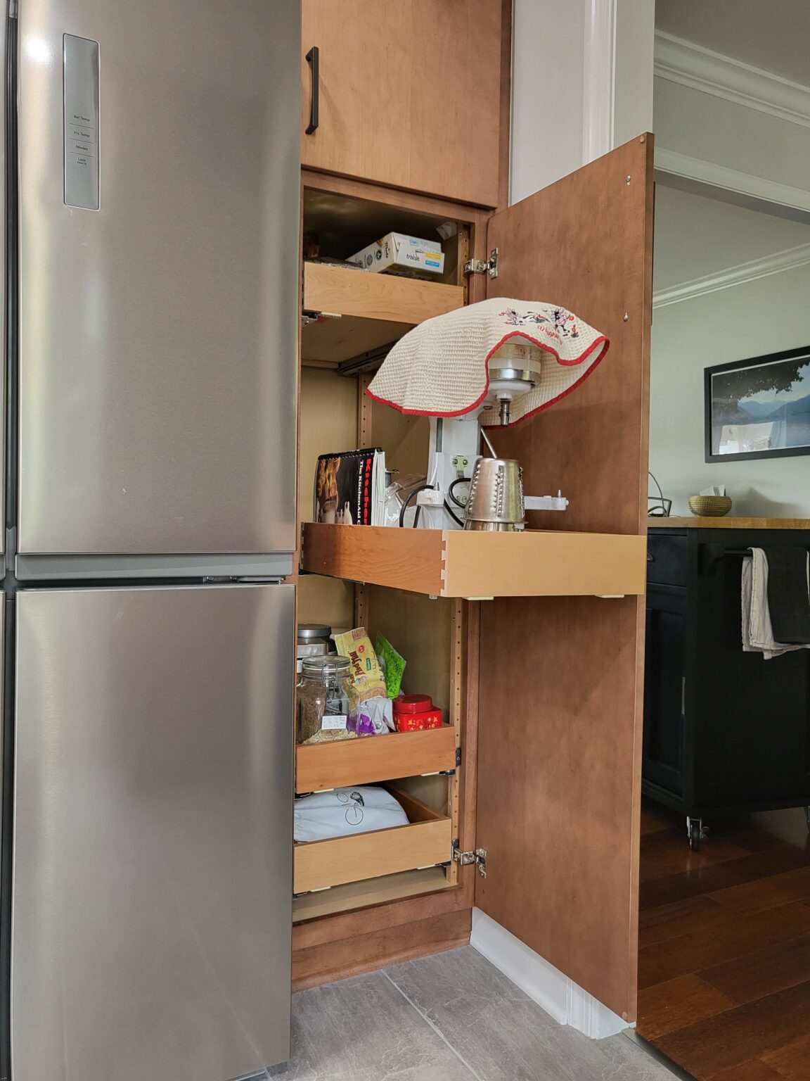 A kitchen cabinet with pull-out shelves holds a covered stand mixer, cookbooks, and various kitchen items next to a stainless steel refrigerator.