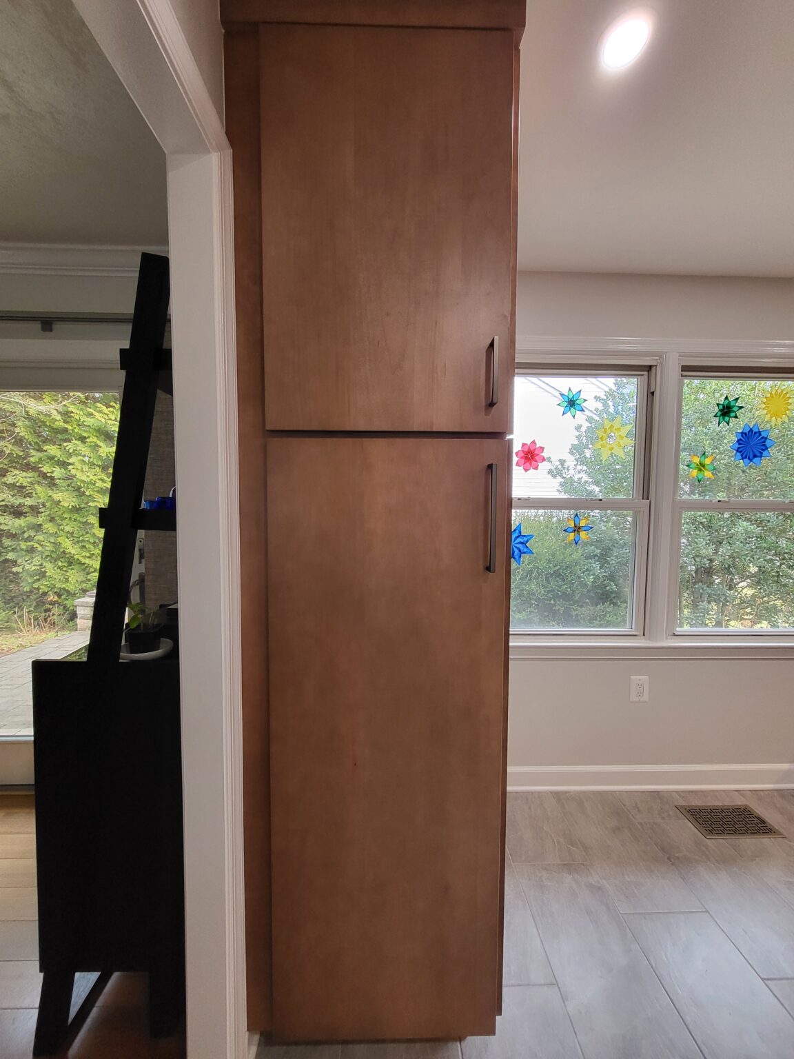 Tall wooden cabinet with two doors stands against a wall near windows decorated with colorful sun-shaped clings. Adjacent rooms are partially visible on either side.