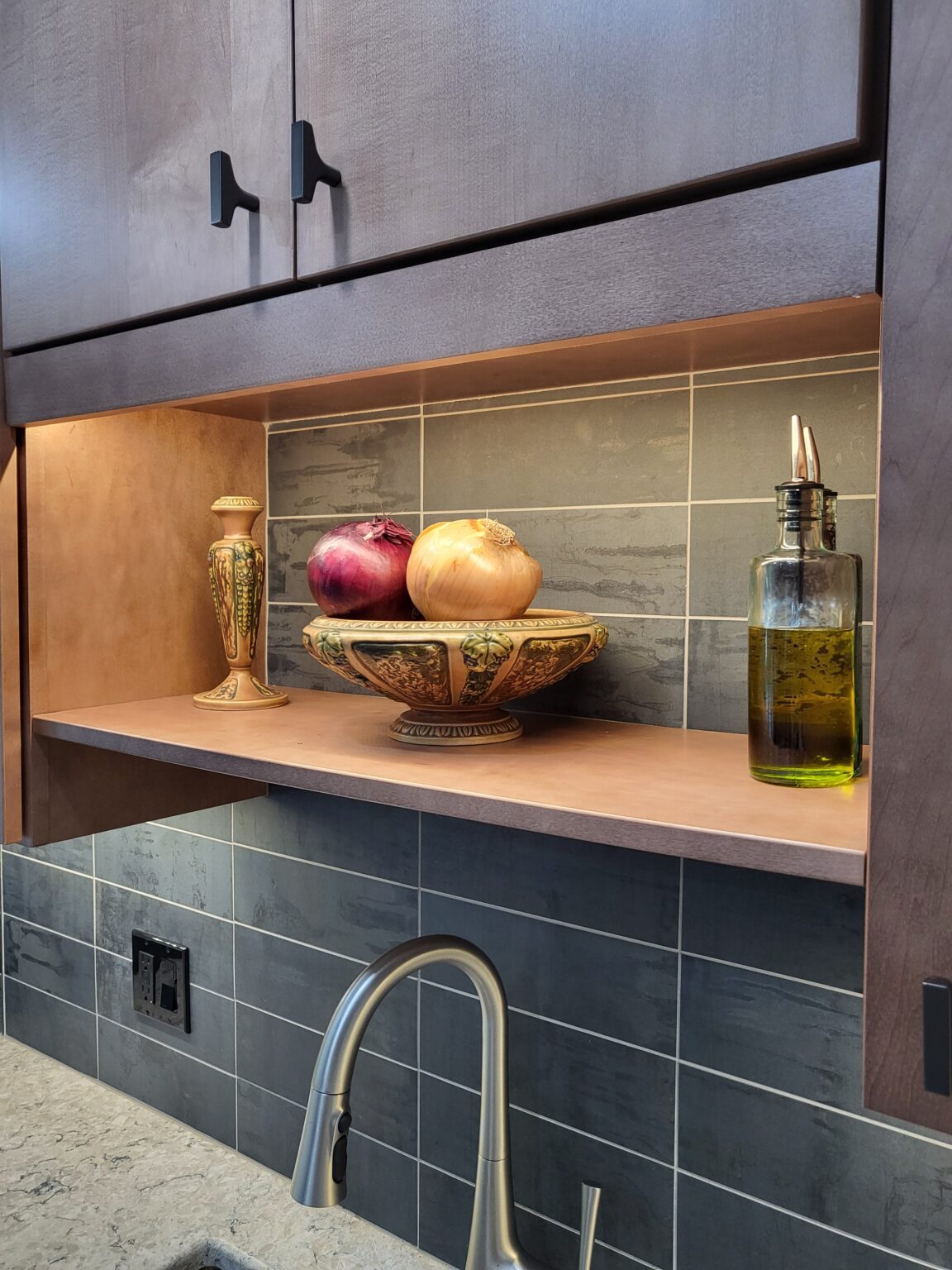 A kitchen shelf displays a decorative bowl with a red and yellow onion, a vase, and a bottle of olive oil above a sink with gray tile backsplash.