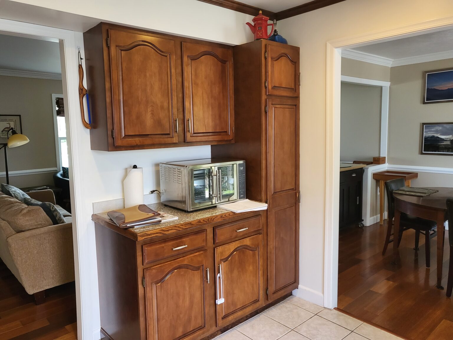 Wooden kitchen cabinets with a countertop holding a microwave, paper towel roll, and some papers; adjoining rooms are visible in the background.