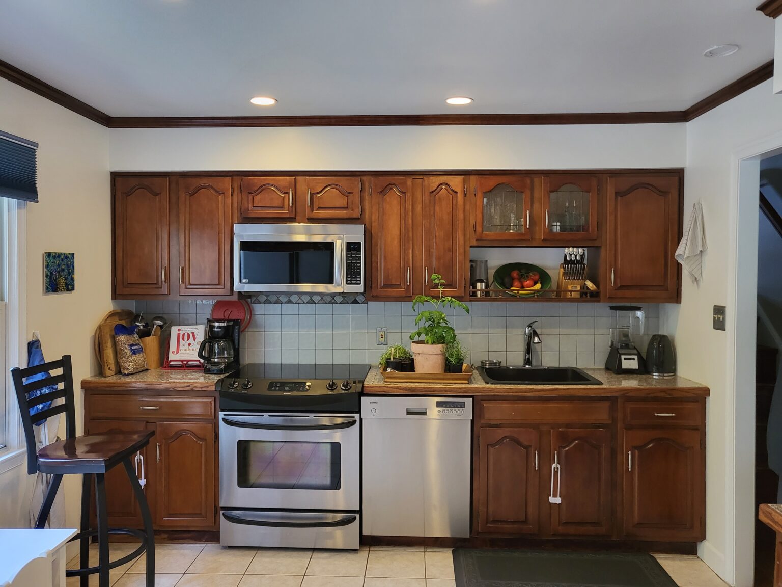 A kitchen with wooden cabinets, stainless steel appliances, a black sink, countertop appliances, a potted plant, and a bar stool, all under recessed lighting.