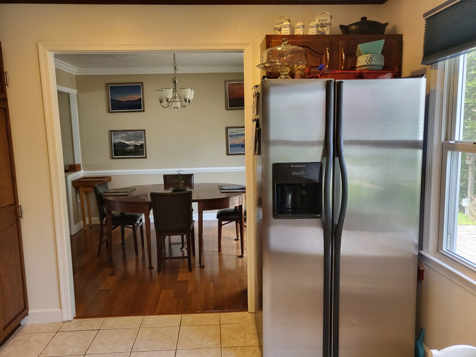 A stainless steel refrigerator stands next to a window in a kitchen, facing an adjoining dining room with a table, chairs, chandelier, and framed photos on the wall.