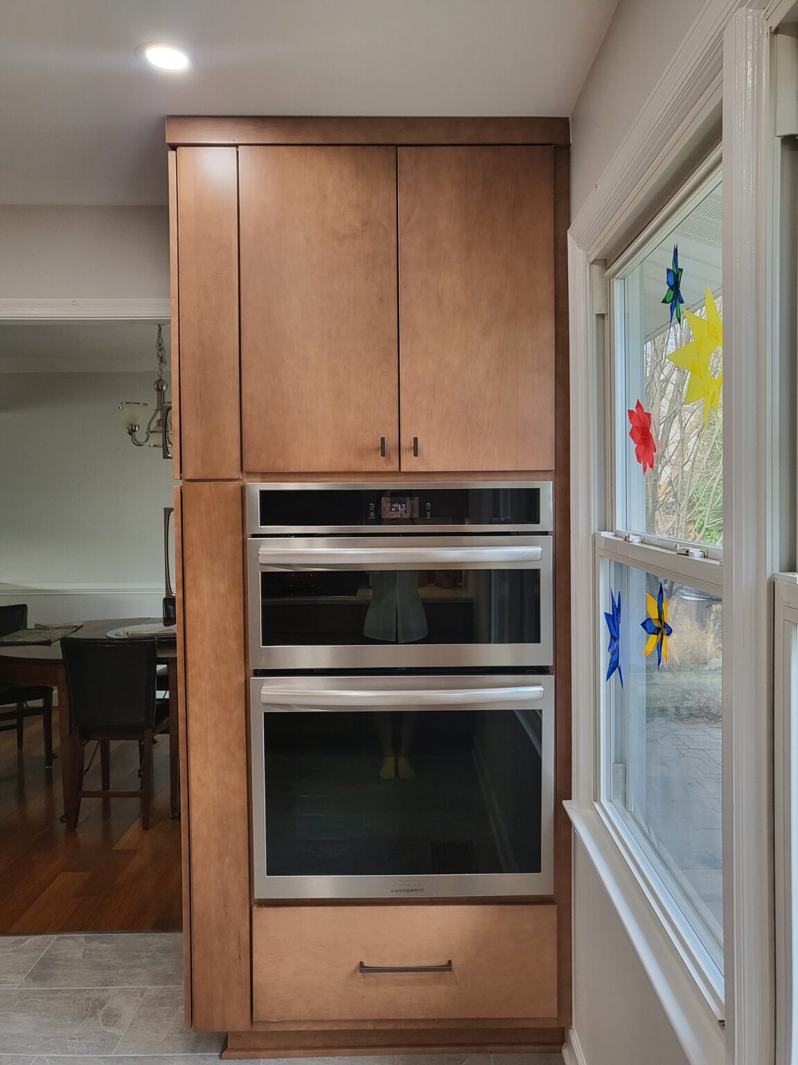 Built-in double wall oven with wood cabinetry next to a window decorated with colorful paper stars; dining area visible in background.