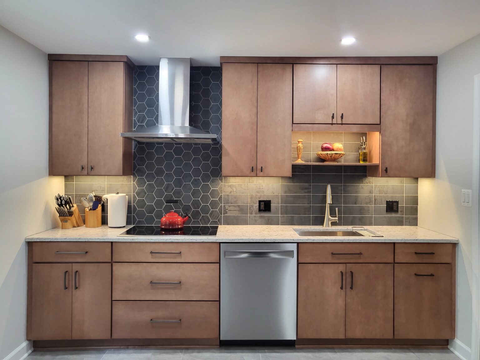 Modern kitchen with wooden cabinets, stainless steel appliances, hexagonal tile backsplash, under-cabinet lighting, and a countertop with utensils and a red teapot on the stove.