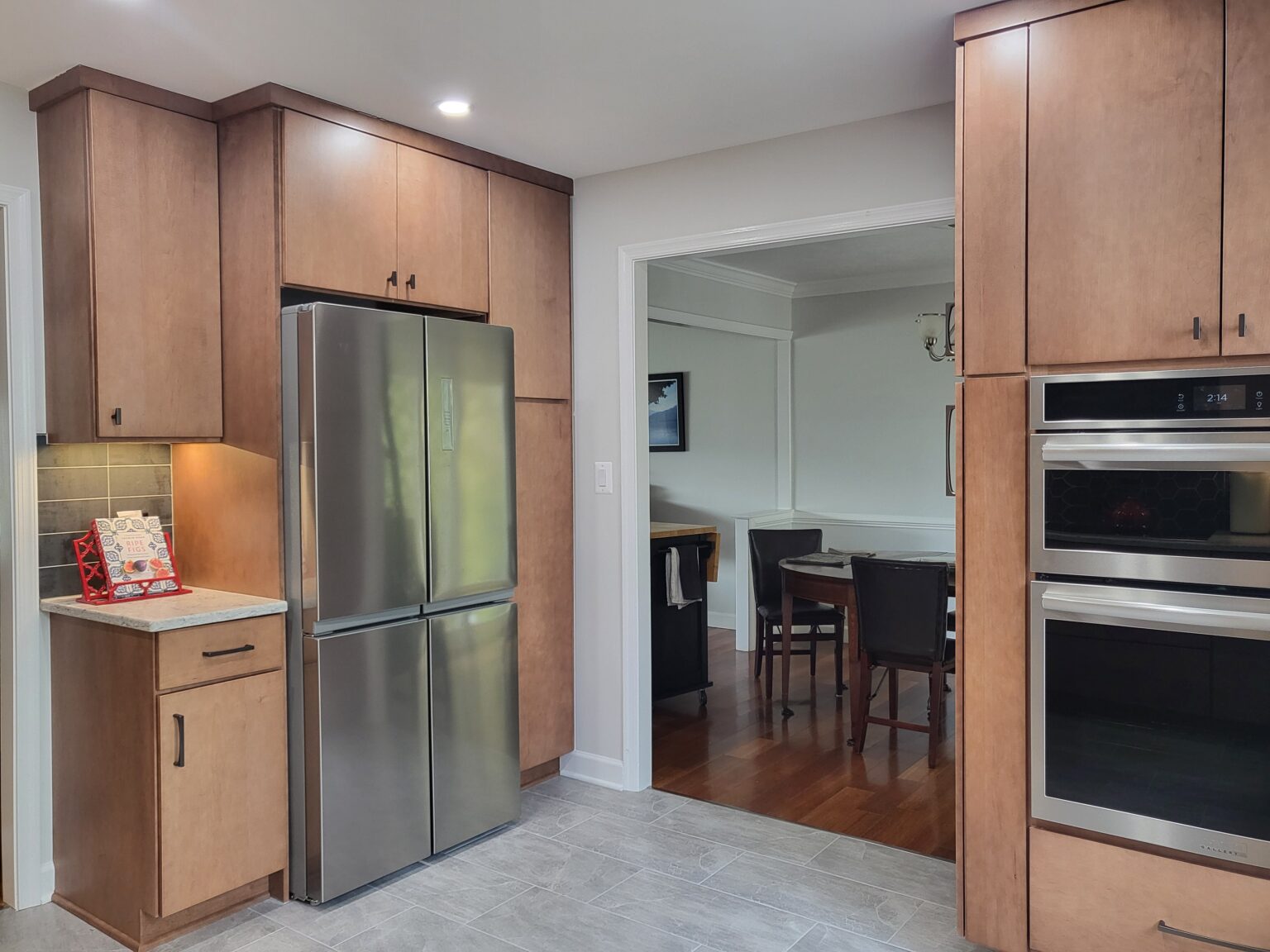 Modern kitchen with light wood cabinets, stainless steel refrigerator, built-in double ovens, and a view into a dining room with dark wood furniture.