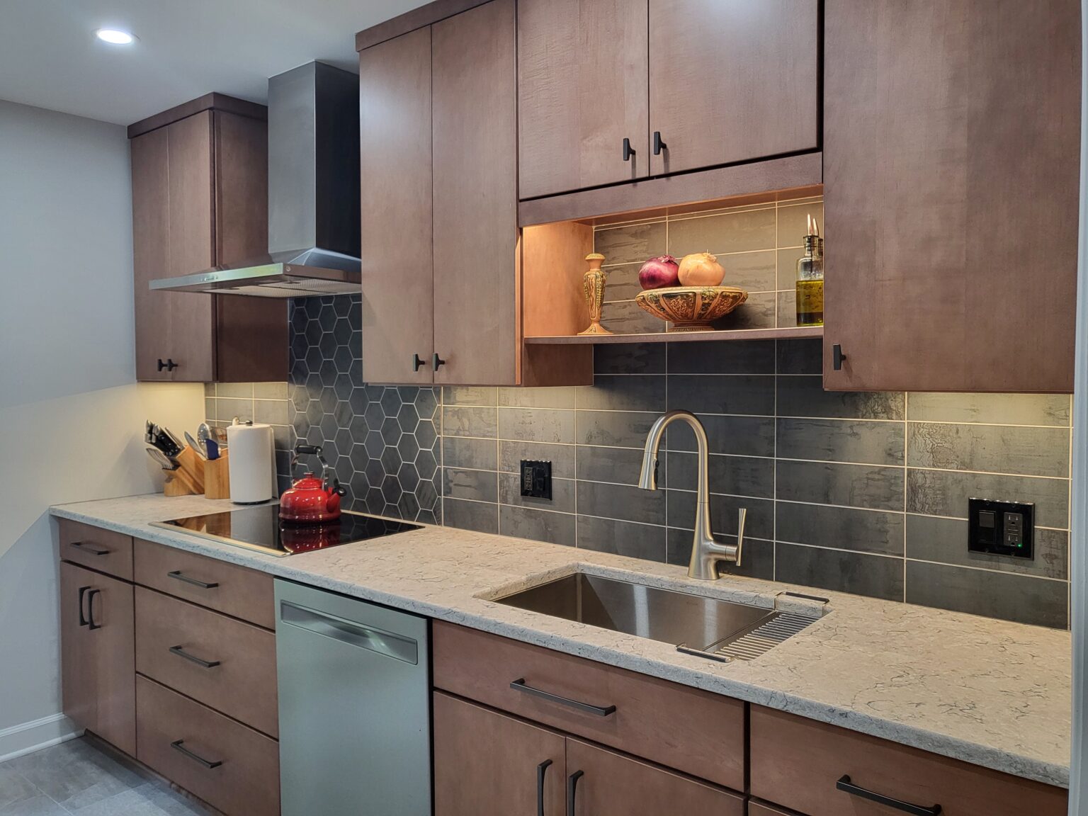 Modern kitchen with wood cabinets, gray tile backsplash, stainless steel appliances, electric cooktop, and a sink under a shelf displaying fruit and bottles.