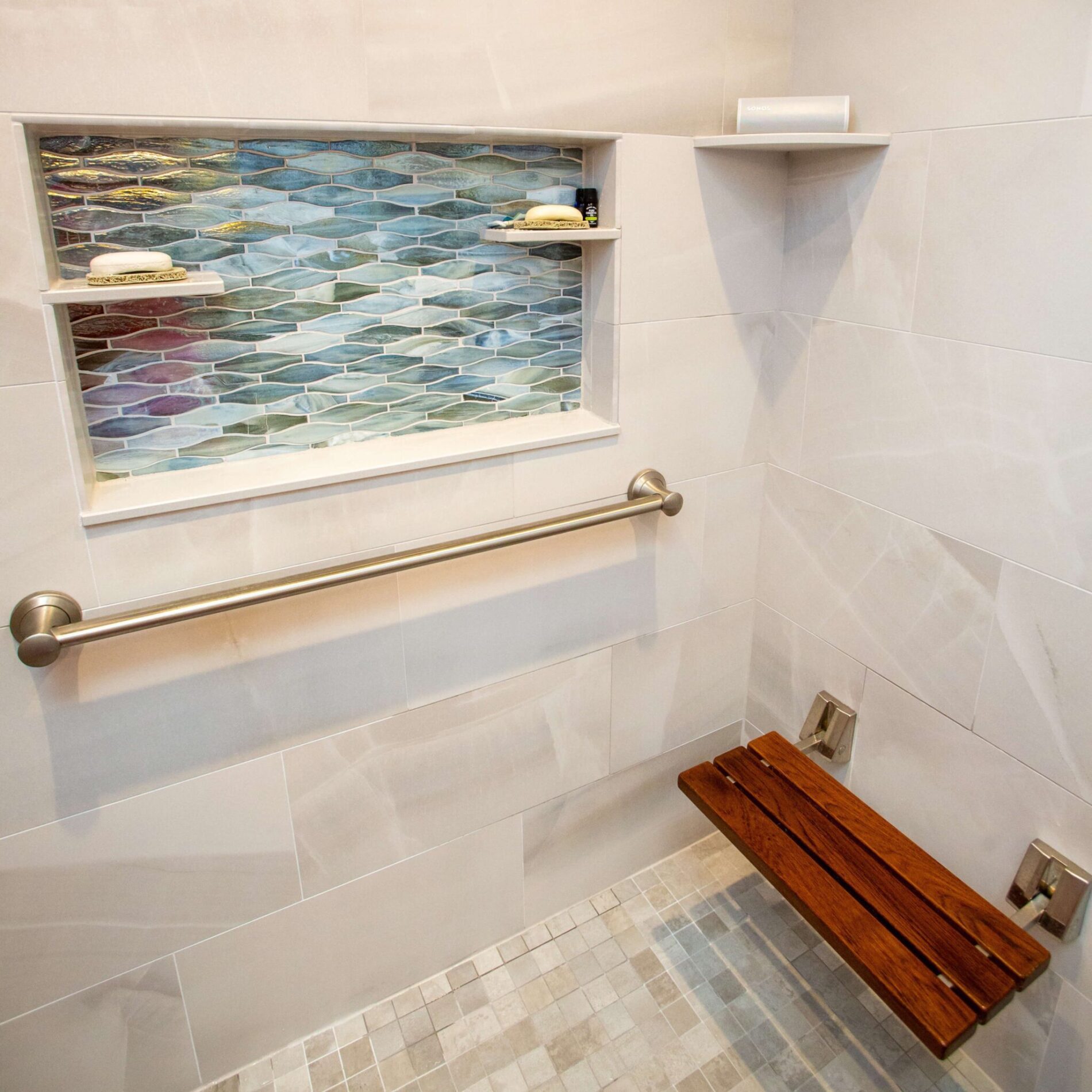 Shower with a wall-mounted grab bar, foldable wooden seat, recessed shelf with blue-green tiles, and soap on shelves—perfect for home remodeling preparedness and enhanced safety.