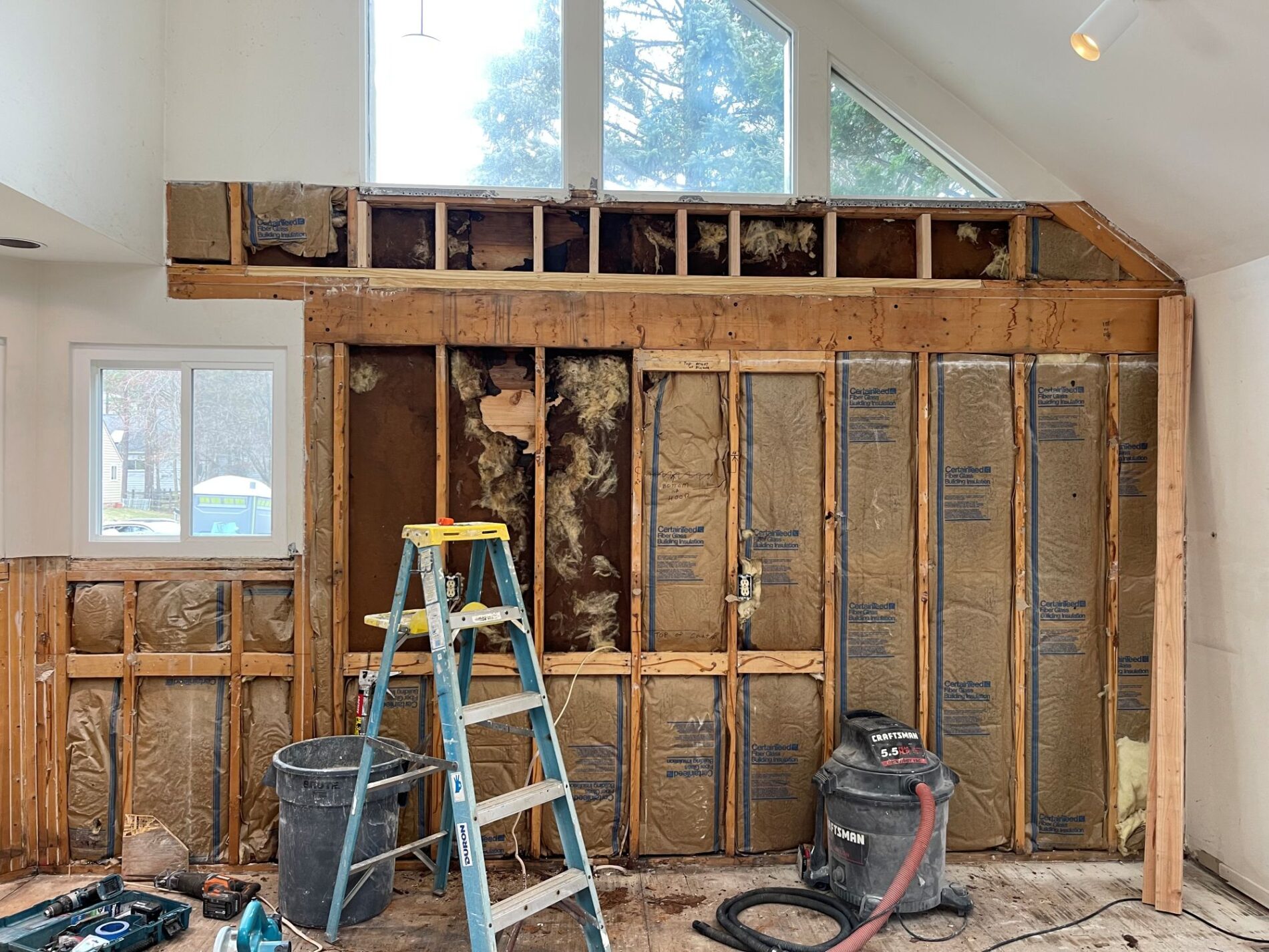 Room under renovation with exposed wall framing and insulation, a ladder, construction tools, vacuum, and debris on the floor highlights the importance of home remodeling preparedness; windows visible in background.