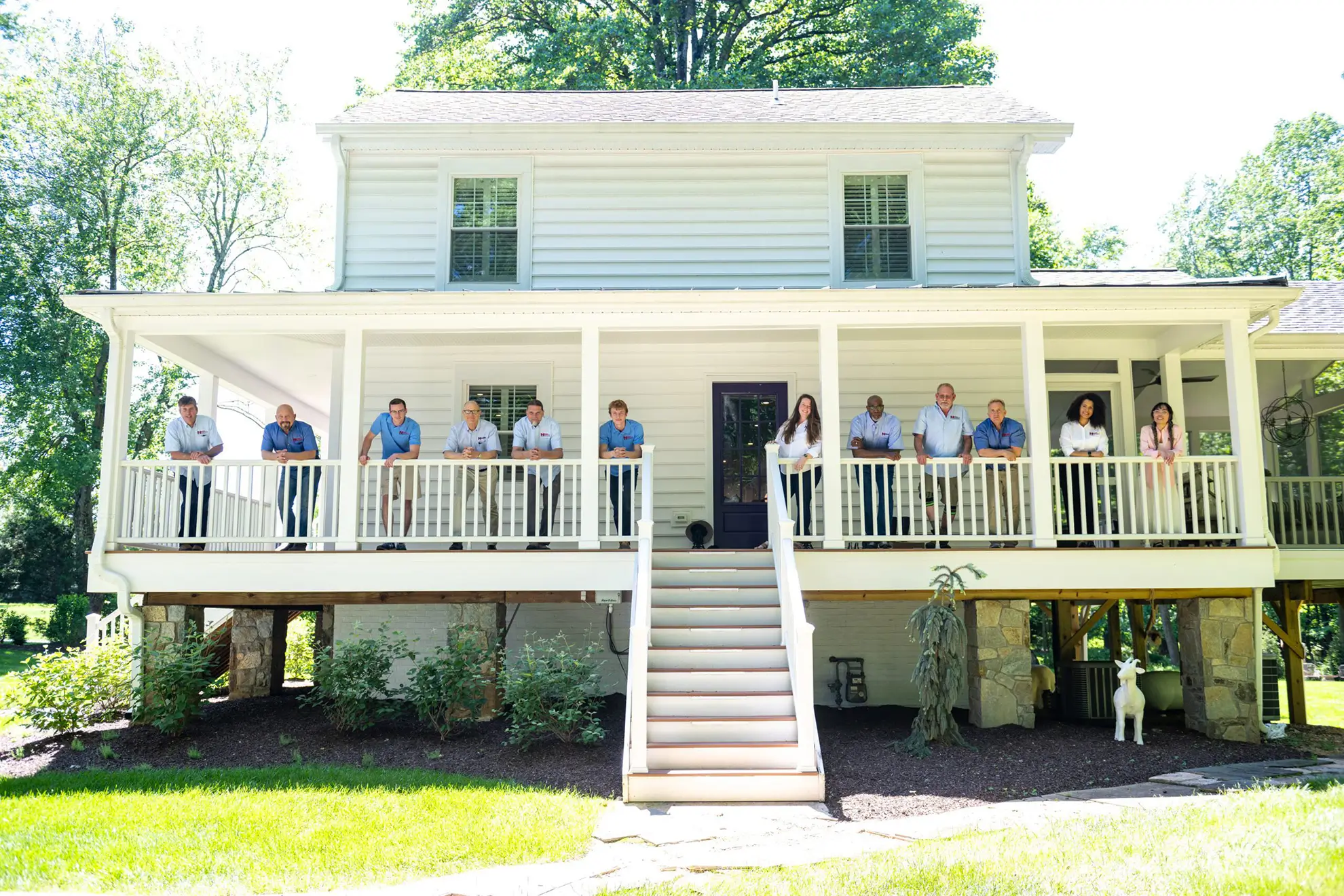 Ten people stand spaced apart along the front porch of a two-story white house on a sunny day, with green trees and grass in the background—perhaps admiring the work of a skilled home remodeling contractor.