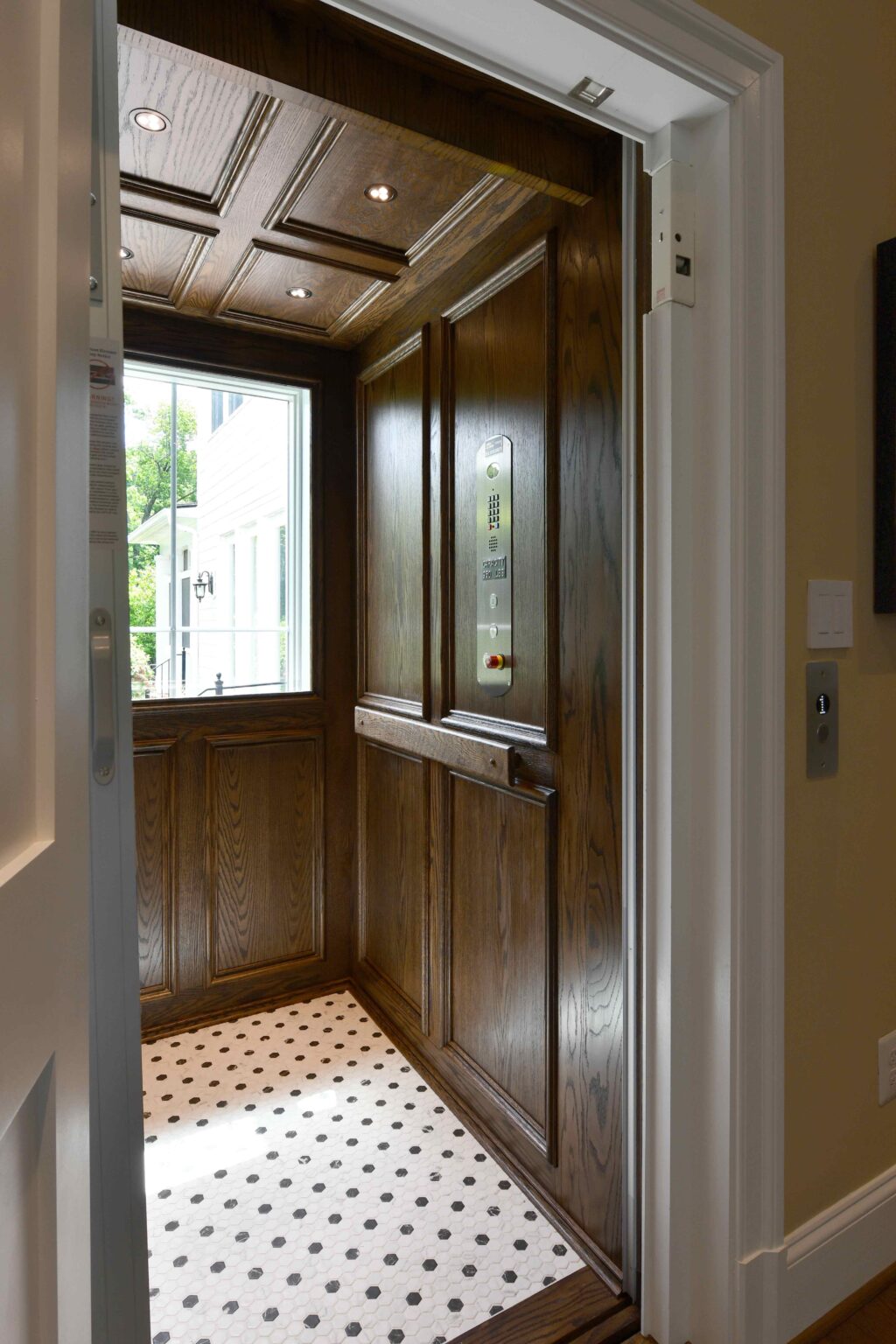 A residential elevator with dark wood paneling, a patterned tile floor, control panel, and window letting in natural light.