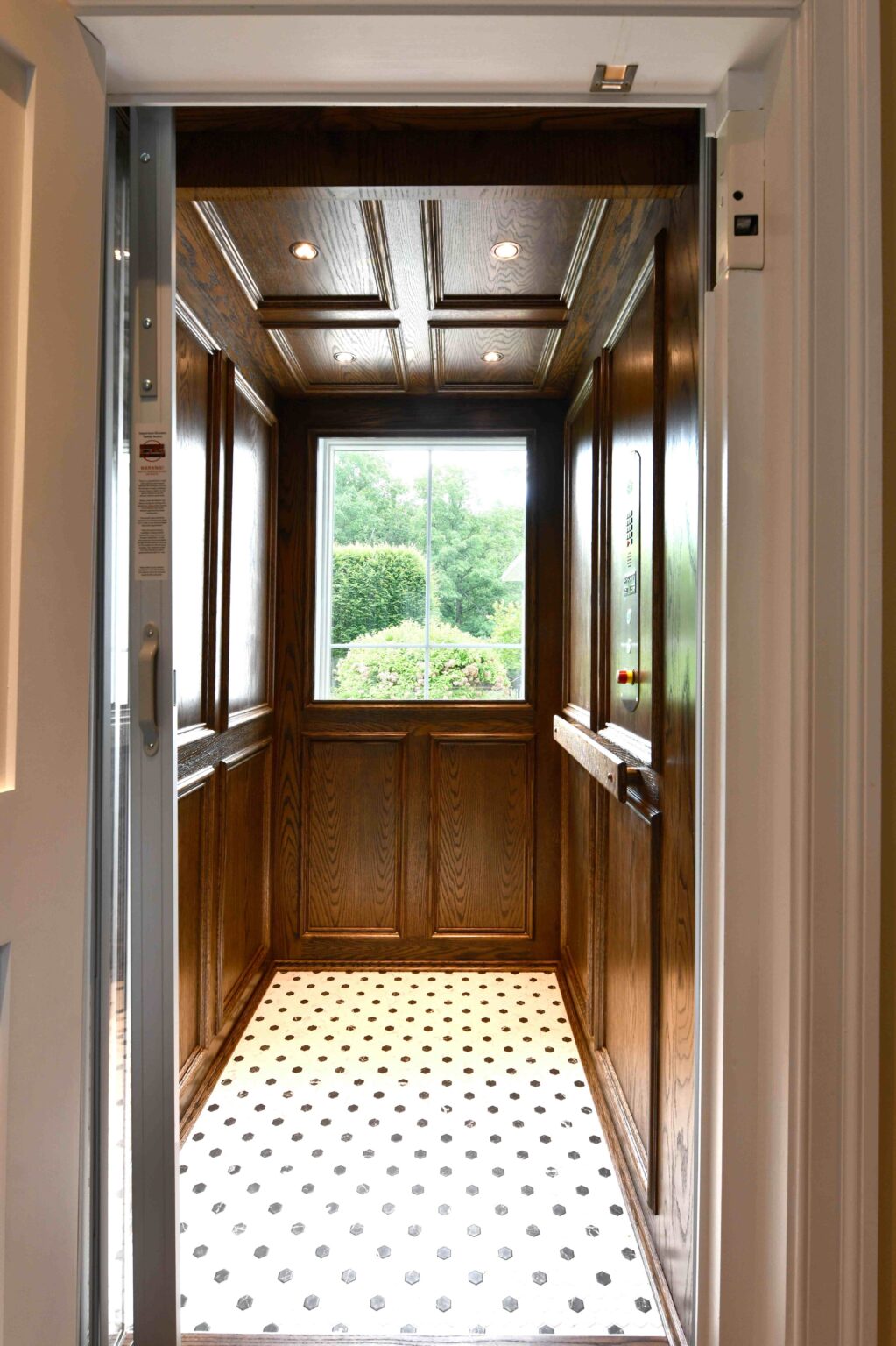 A small wood-paneled residential elevator with a patterned tile floor and a window showing greenery outside.