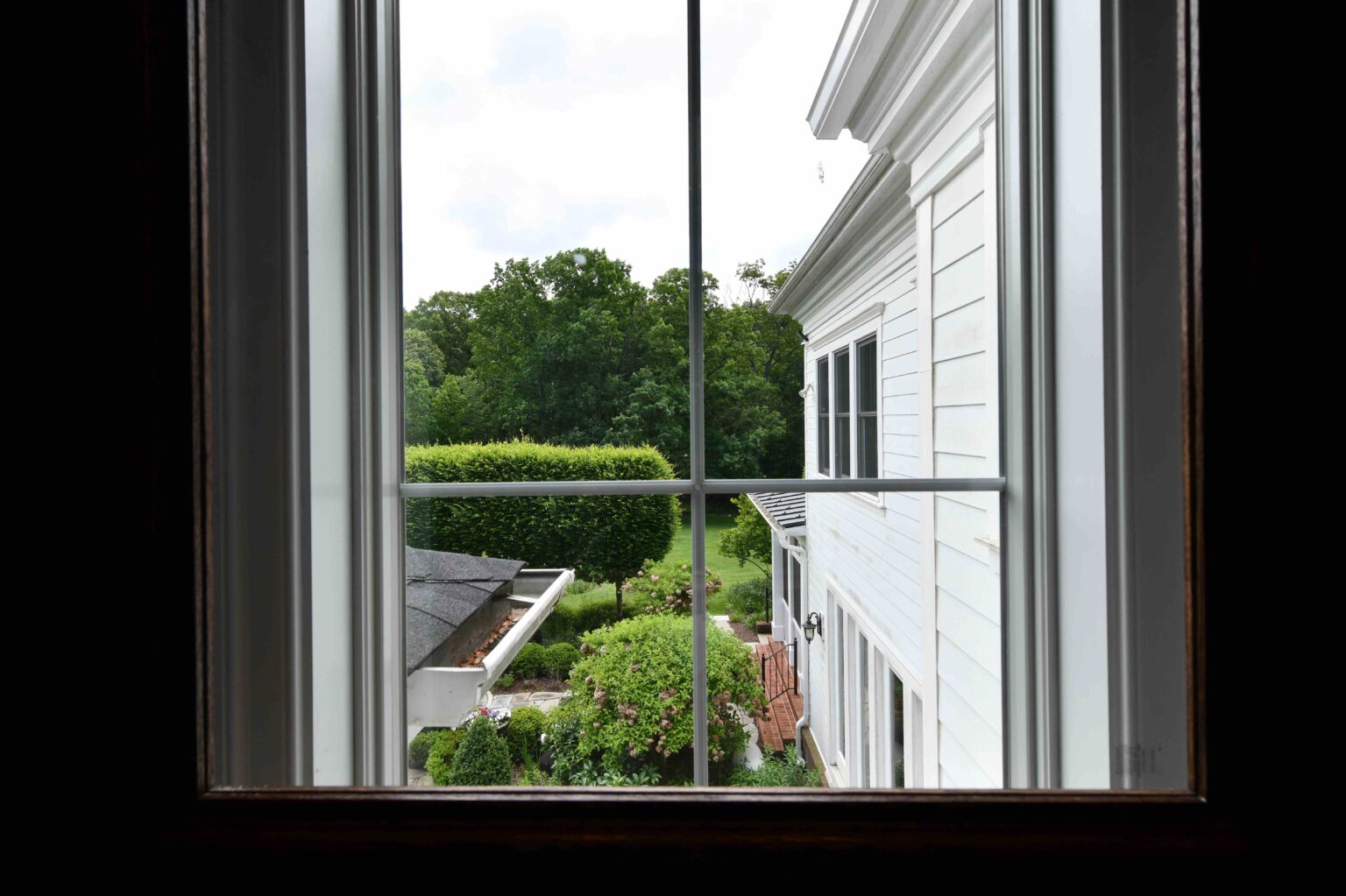 View through a window showing a white building, greenery, trees, and part of a roof outside under a cloudy sky.