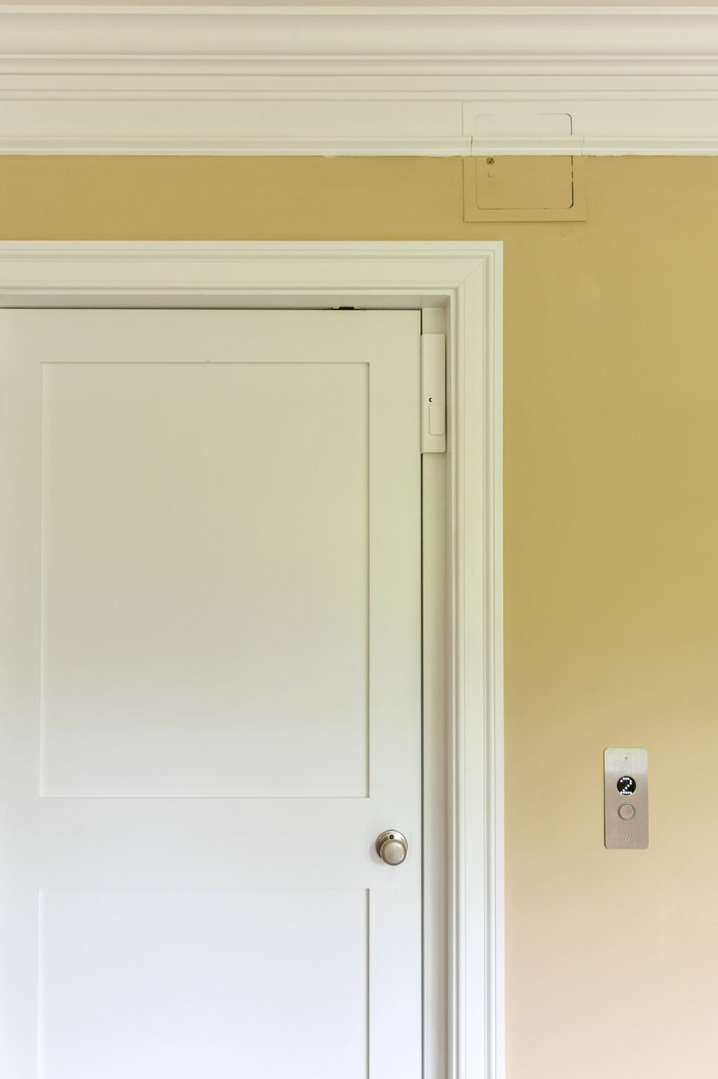 A white door with a silver knob set in a beige wall; to the right, an elevator call button panel is mounted on the wall.