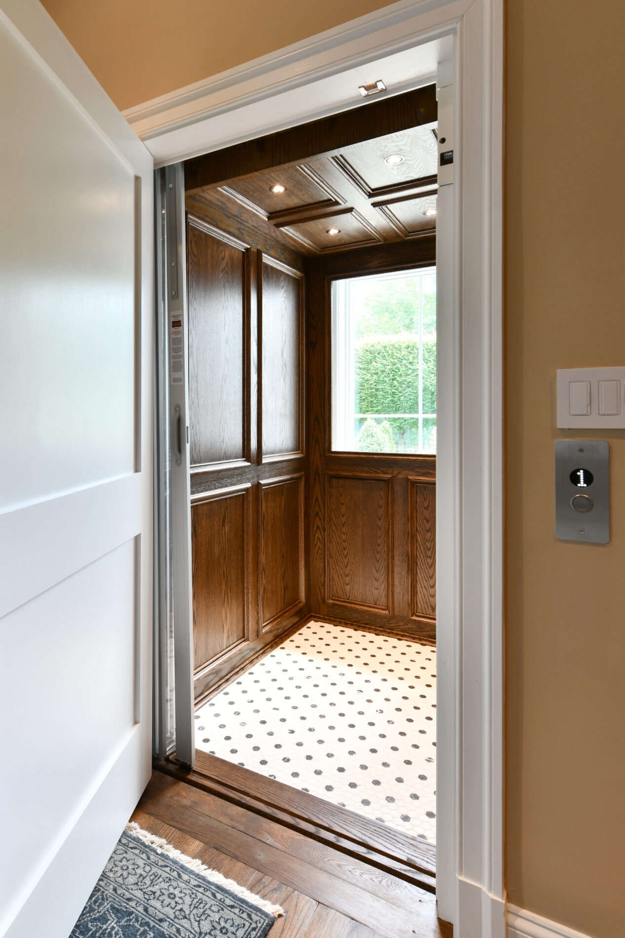 Residential elevator with wood paneling, white and black tile floor, open door, and a window letting in natural light. Control panel with floor button is visible on the wall outside.
