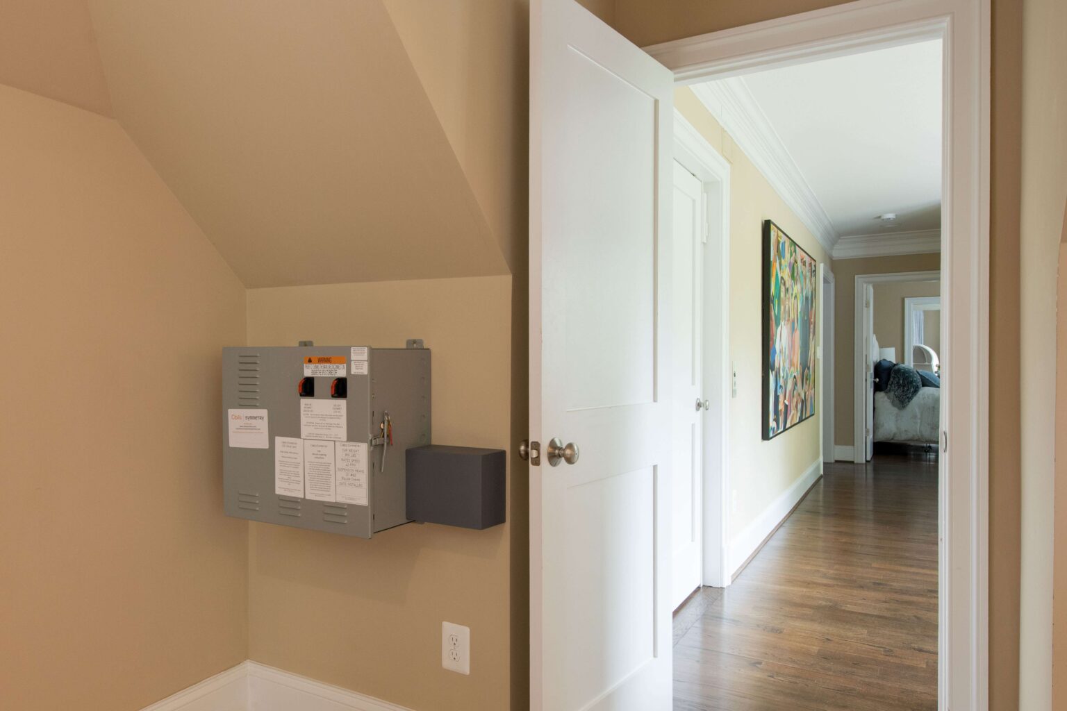A gray electrical panel is mounted on a beige wall in a hallway with hardwood floors and an open white door leading to a brightly lit room.
