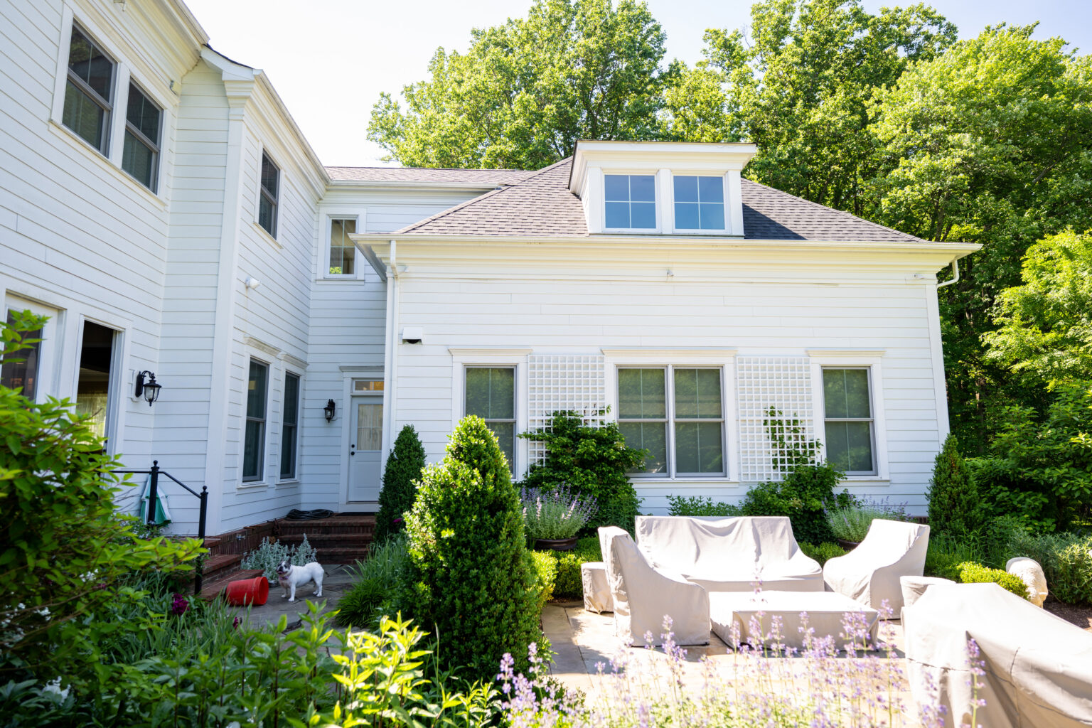 White two-story house with a small patio featuring outdoor seating, surrounded by greenery and flowering plants on a sunny day.