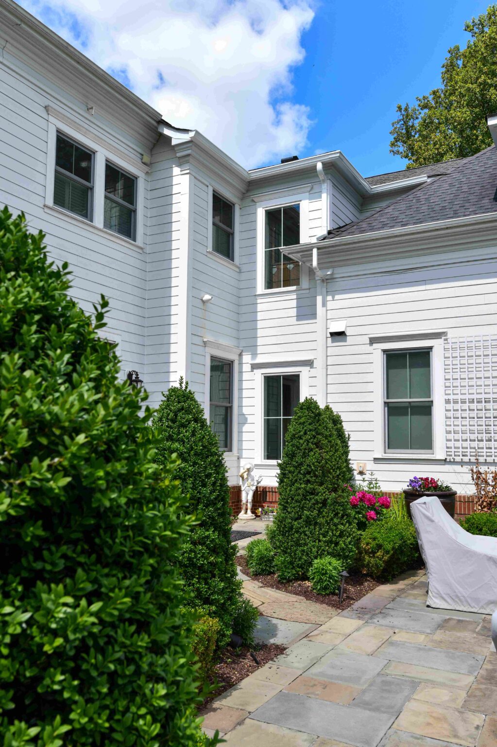 A white two-story house with several windows overlooks a patio area, landscaped with trimmed bushes, flowering plants, and stone pavers under a partly cloudy sky.