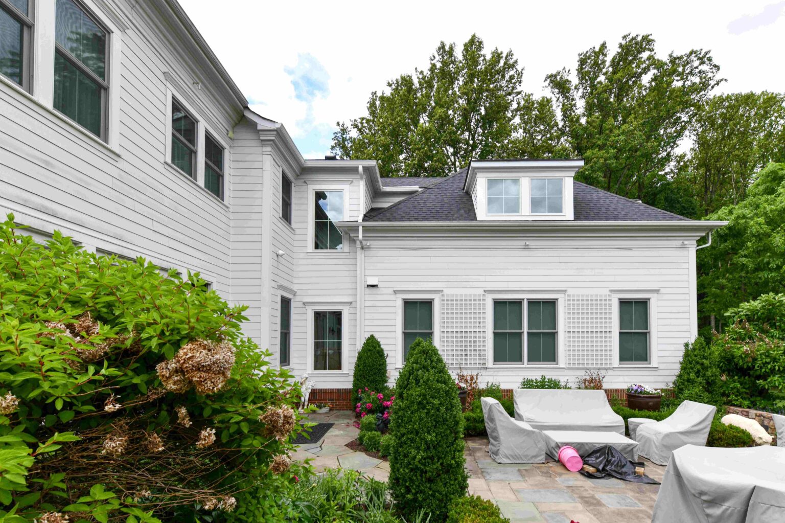 A white two-story house with green-trimmed windows, patio furniture covered with tarps, and bushes lining a stone patio.