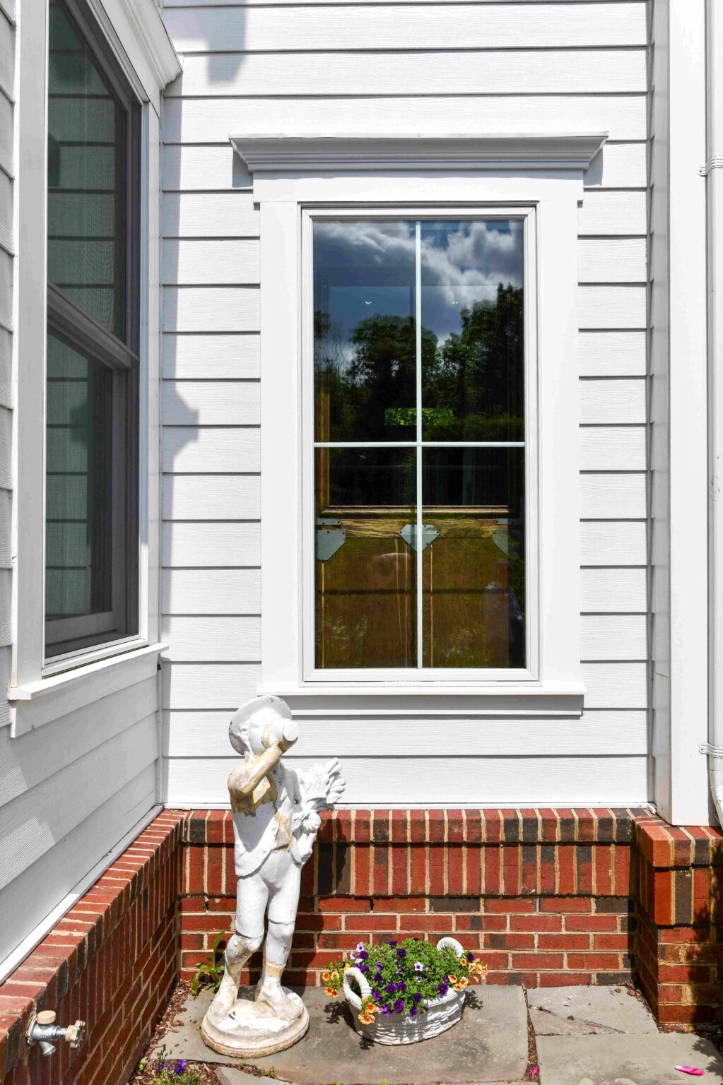 A white statue of a child with a bird stands next to a pot of flowers beneath a window on a white siding house with red brick trim.