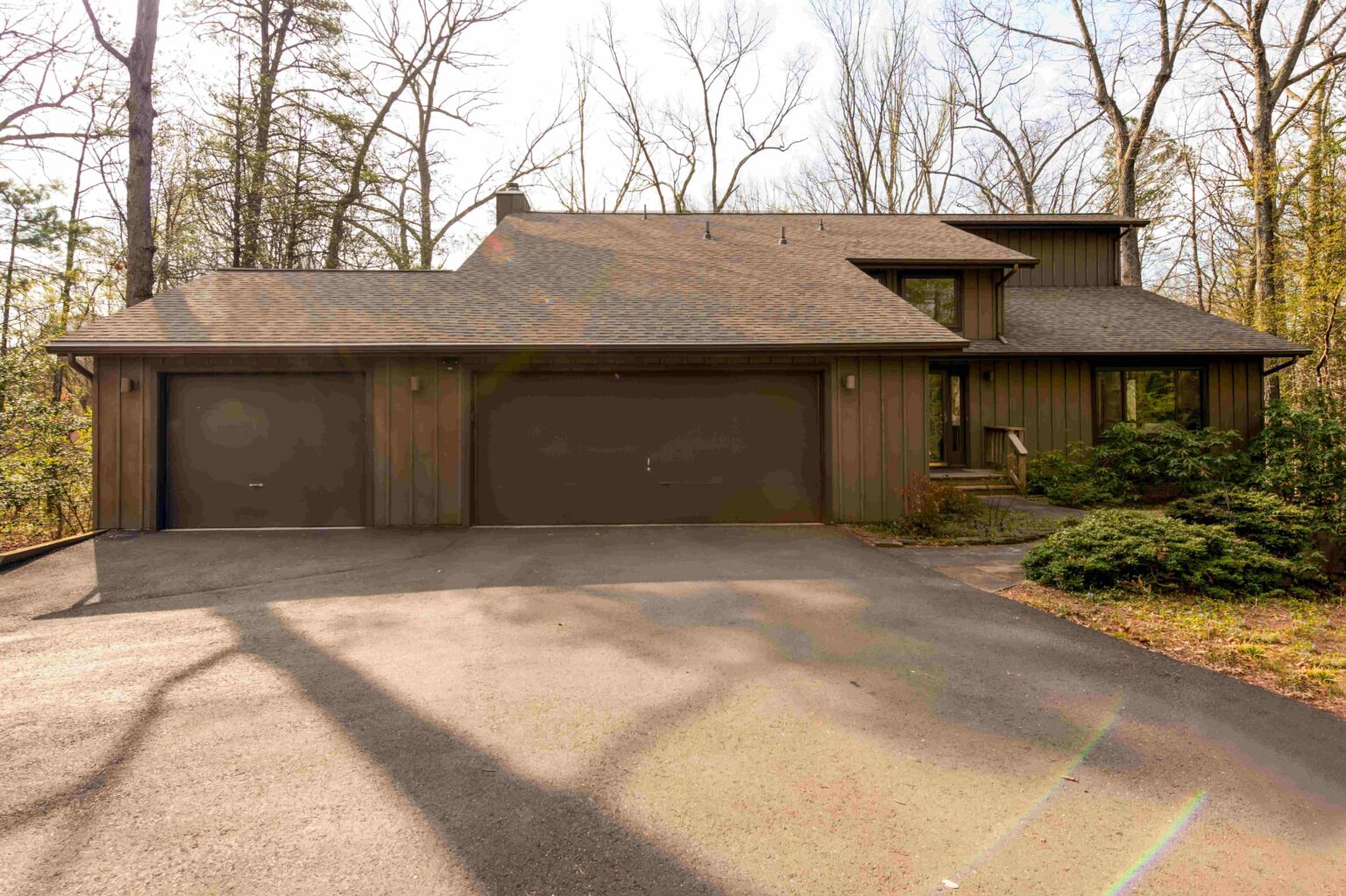 Two-story brown house with a three-car garage, surrounded by trees and a paved driveway, under clear daylight.