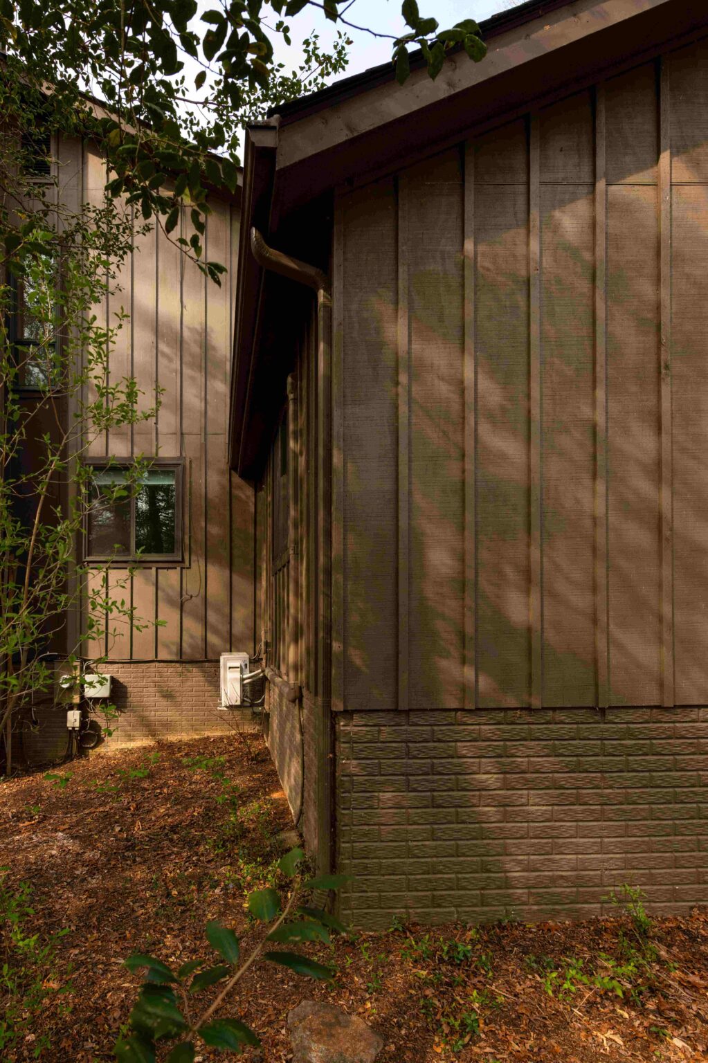 Exterior corner of a brown house with vertical siding and a brick foundation, surrounded by dirt, leaves, and a few green plants in the foreground.