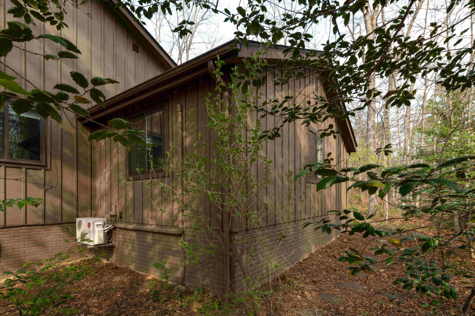 A wooden house exterior with a small window, corner brick foundation, and a wall-mounted air conditioning unit, surrounded by trees and leafy vegetation.