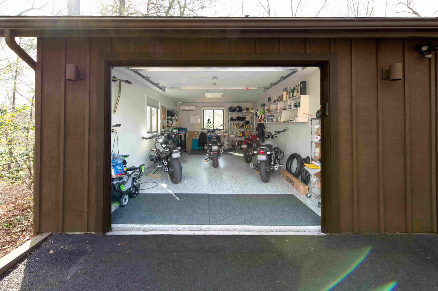 Open garage with three motorcycles parked inside, shelves holding tools, tires, and boxes, and a window at the back letting in natural light.