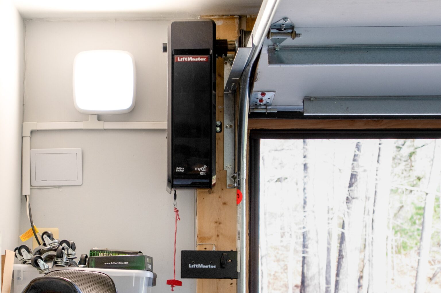 A LiftMaster garage door opener is mounted vertically on a wooden support next to a partially open garage door in a well-lit garage.