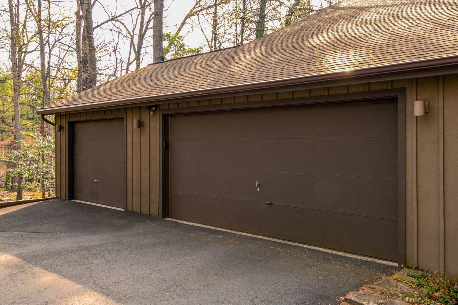 Two closed brown garage doors on a wooden house with a sloped driveway and trees in the background.