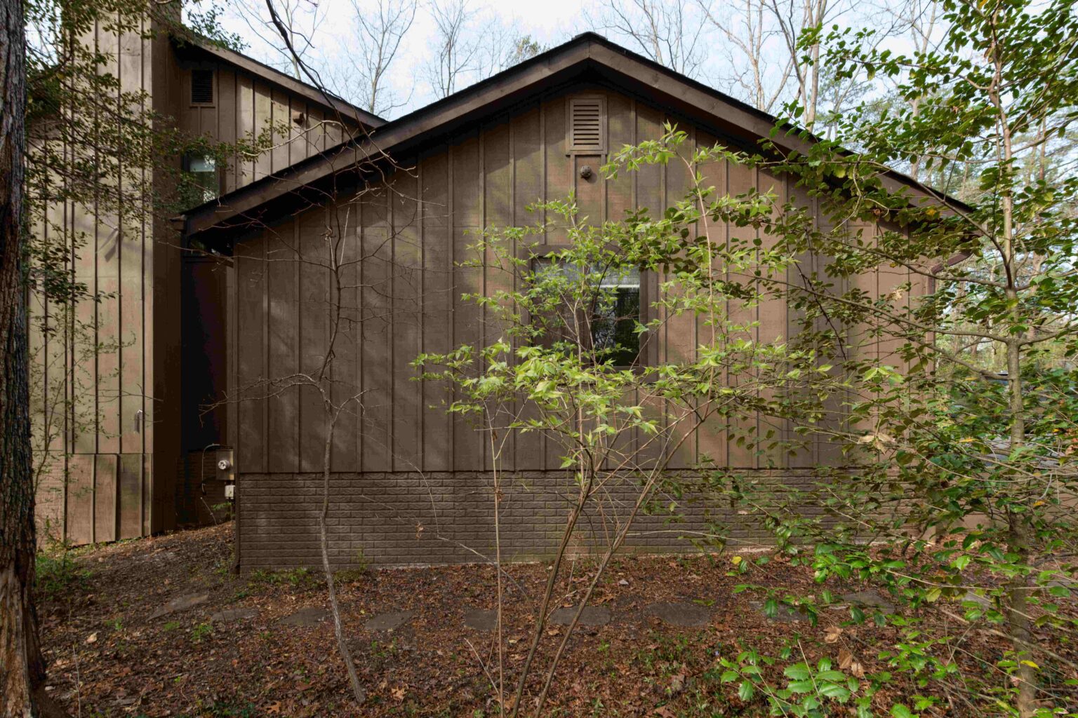 A brown wooden house with a brick foundation, surrounded by trees and dense foliage, viewed from the side.