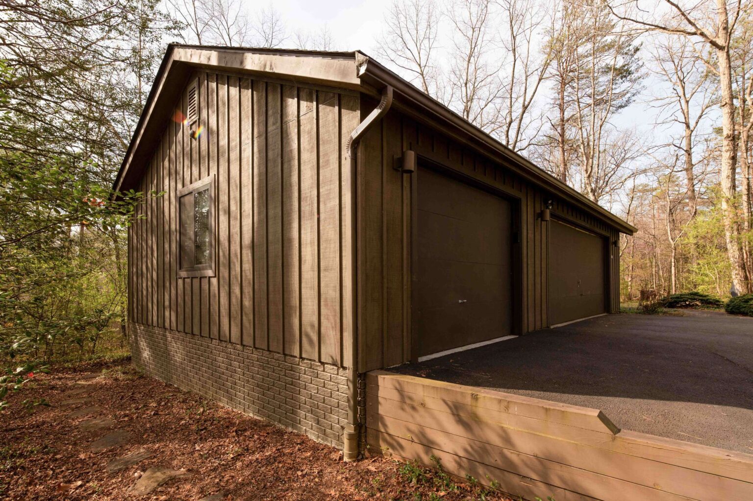 A brown wooden garage with two closed doors, a gable roof, one side window, and a paved driveway, surrounded by trees and fallen leaves.