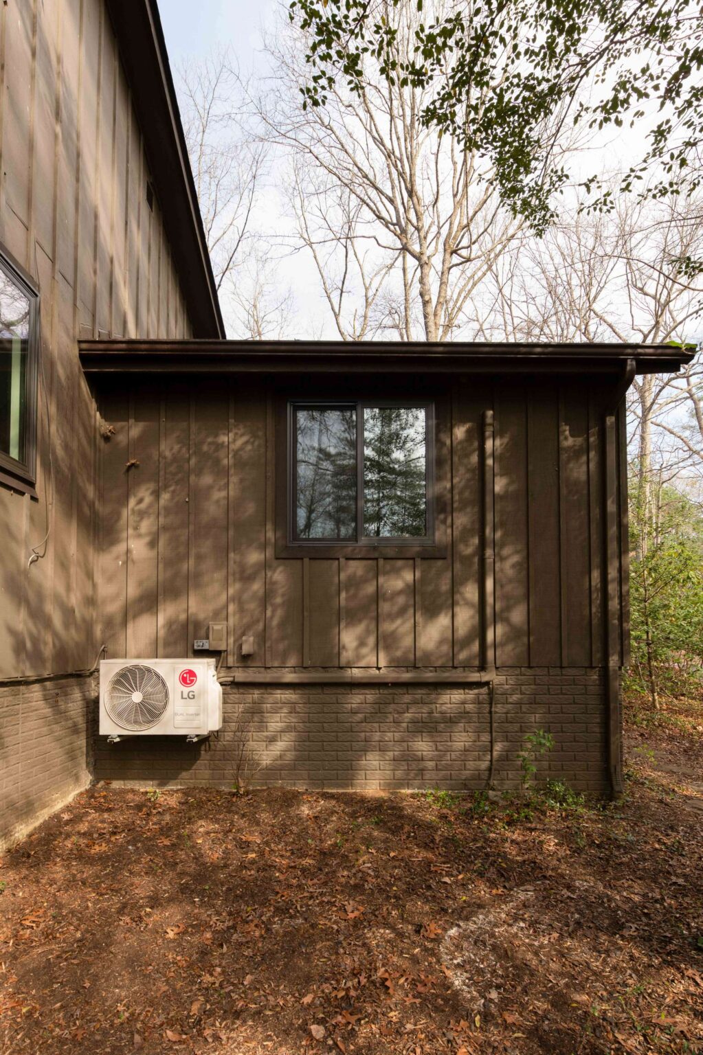 A brown-sided house with a rectangular window and an LG outdoor air conditioning unit mounted on the lower wall, surrounded by bare trees and dry ground.