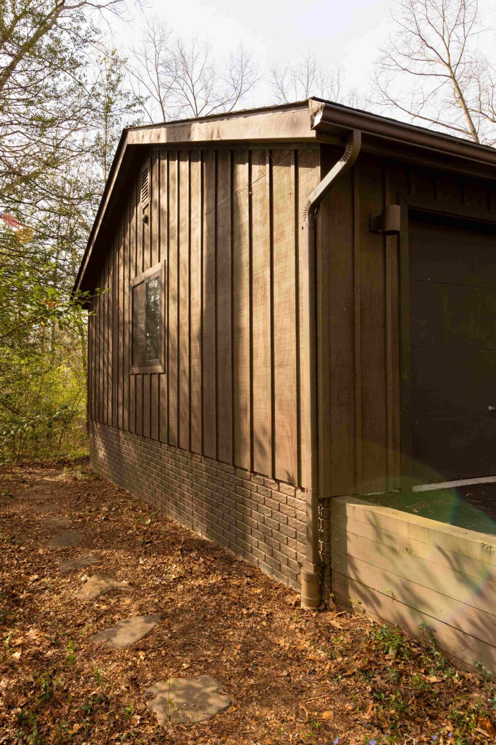 A brown wooden building with vertical siding and a small window, surrounded by trees and dry leaves on the ground.
