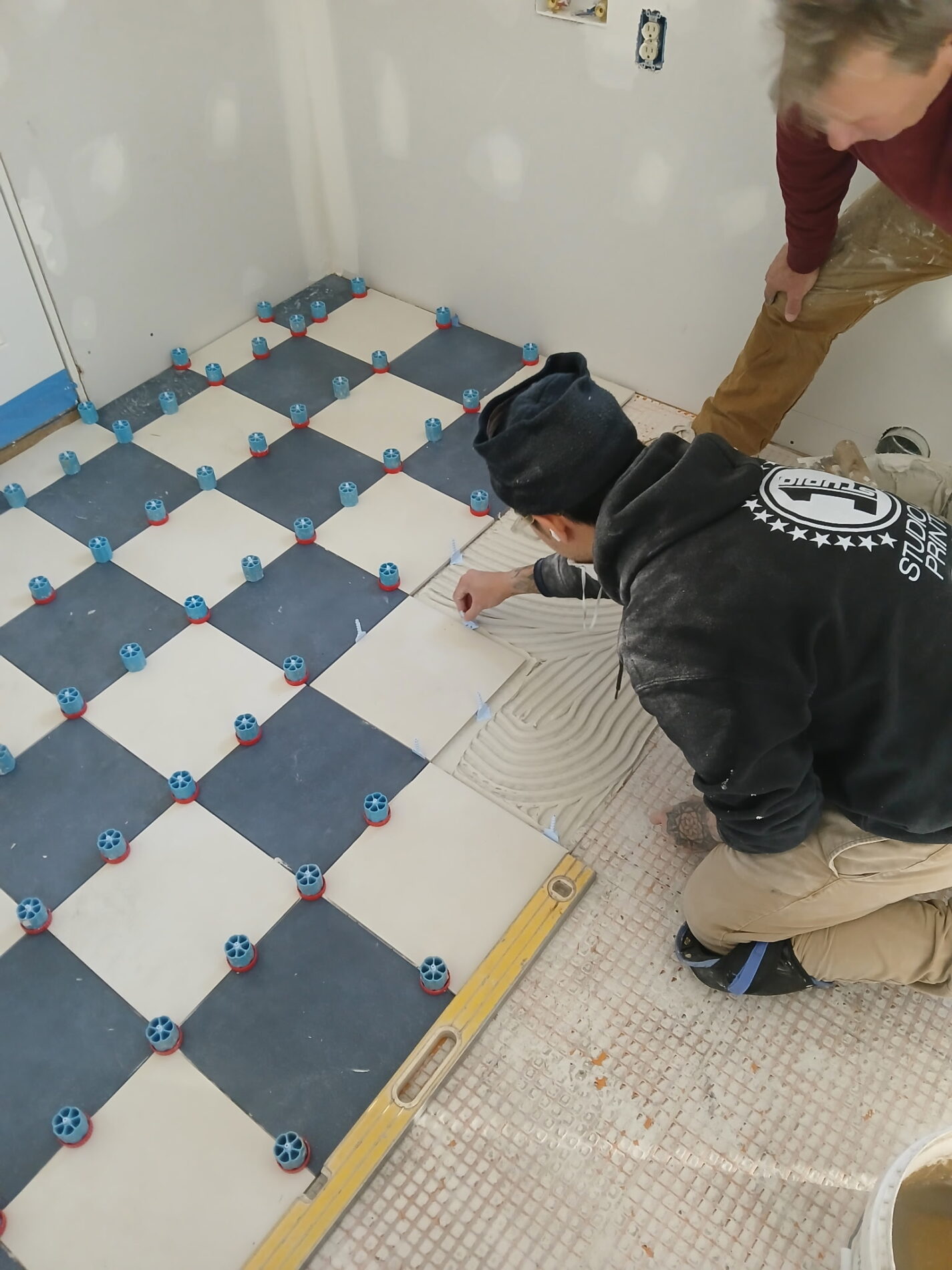 Two people are installing large square tiles on a floor, using spacers for alignment. One person is placing a tile while the other observes.