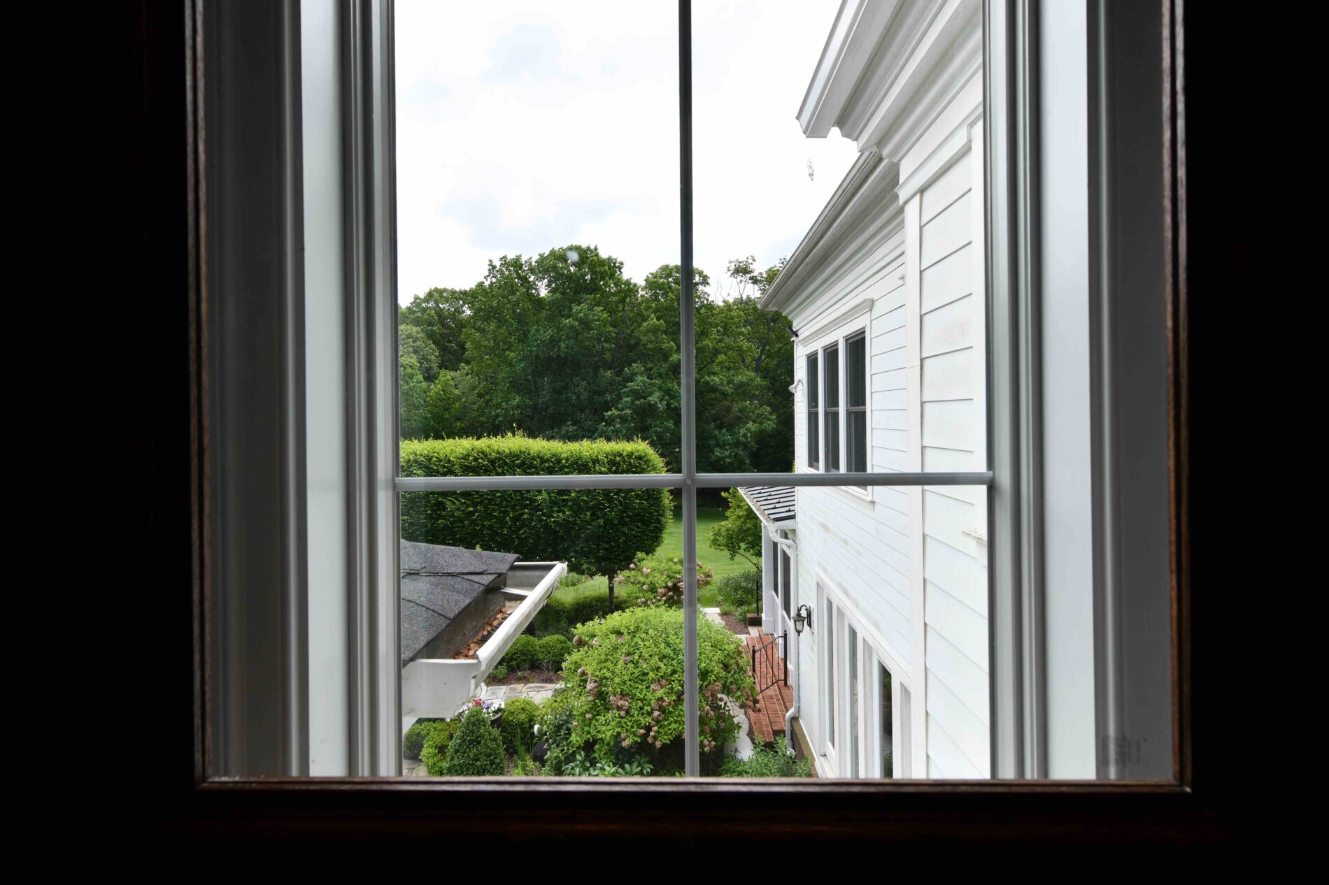 View through a window showing the side of a white house, green trees, manicured bushes, and part of a garage roof in an outdoor garden setting.