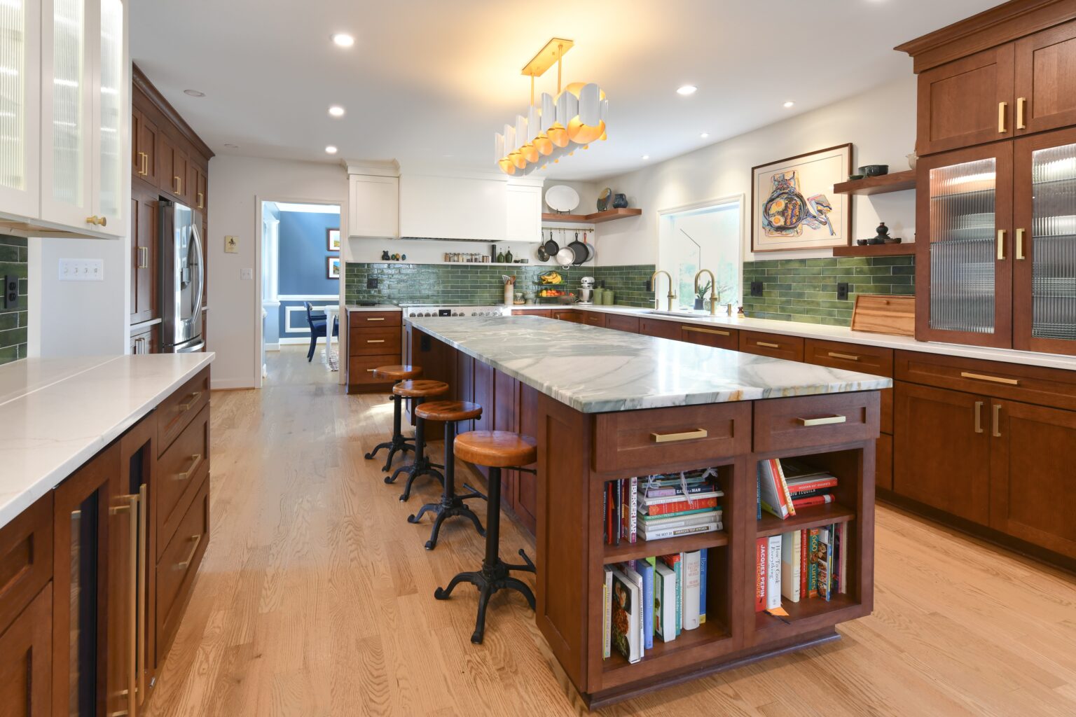 Spacious kitchen with wood cabinets, marble island, green tile backsplash, and bar stools. Books are stored in the island shelves, and modern lighting hangs overhead.