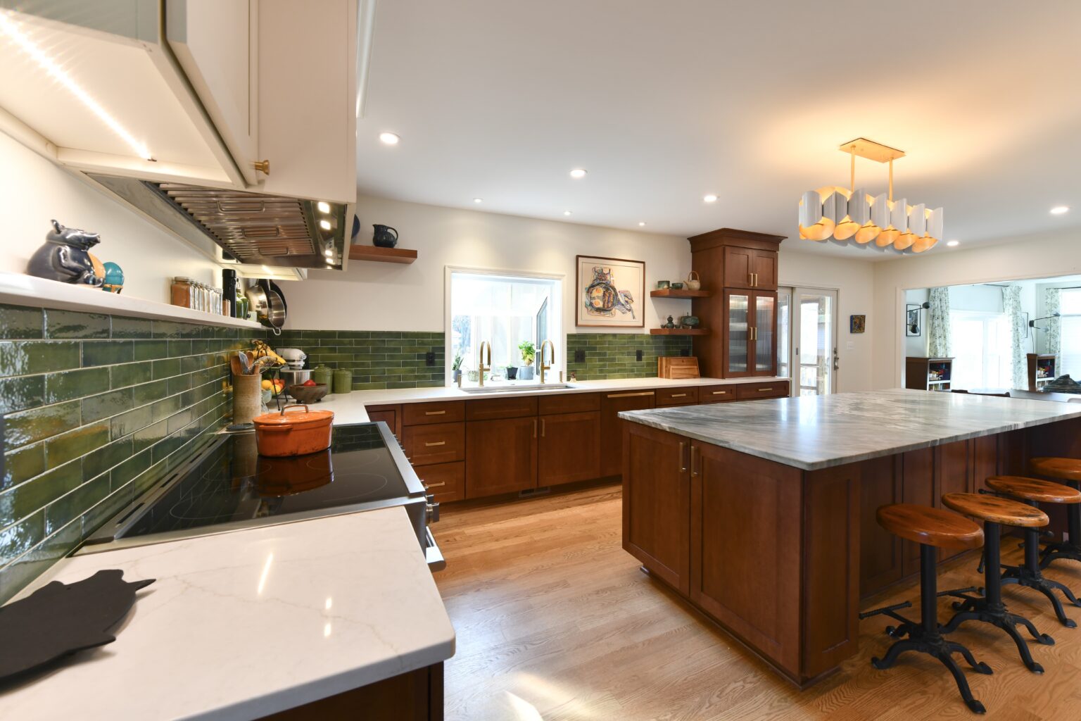 Spacious kitchen with wood cabinets, green tile backsplash, large island with marble countertop, and four wooden stools; sunlight streams through a window above the sink.