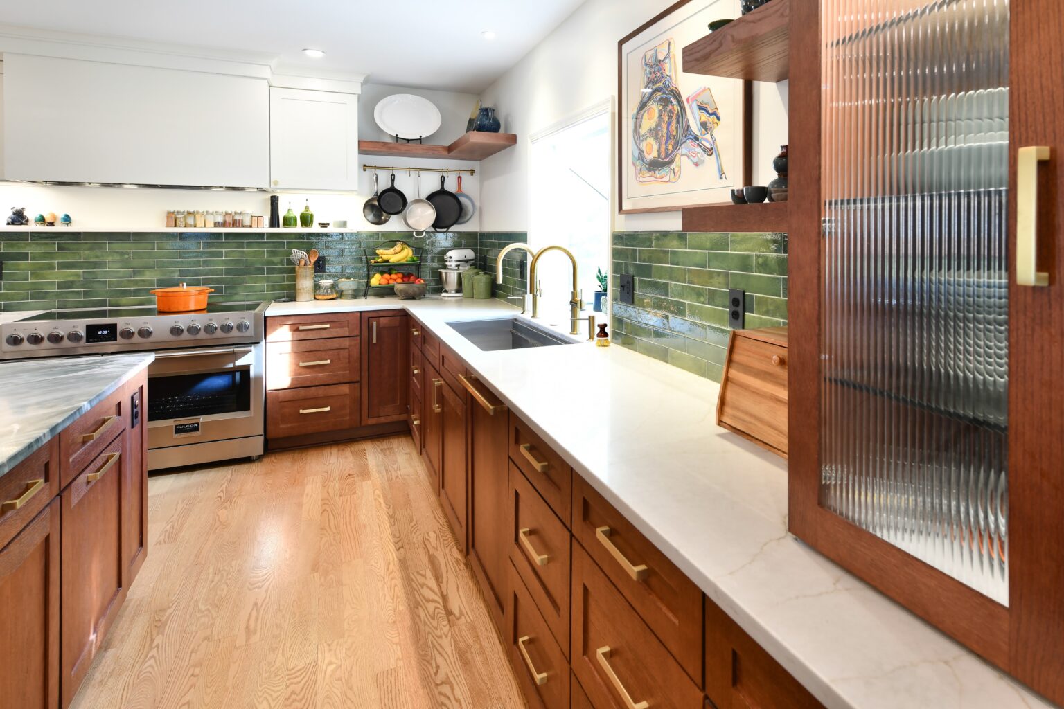 Modern kitchen with wood cabinets, green tile backsplash, marble countertops, stainless steel stove, and various kitchen items on counters and shelves. Natural light enters from a window.