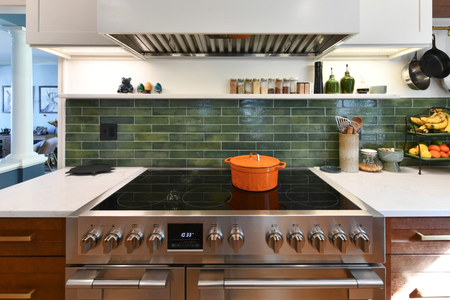 Modern kitchen stovetop with an orange pot on a black glass cooktop, green tiled backsplash, spices, fruit, and kitchenware arranged neatly on the counter and shelves.
