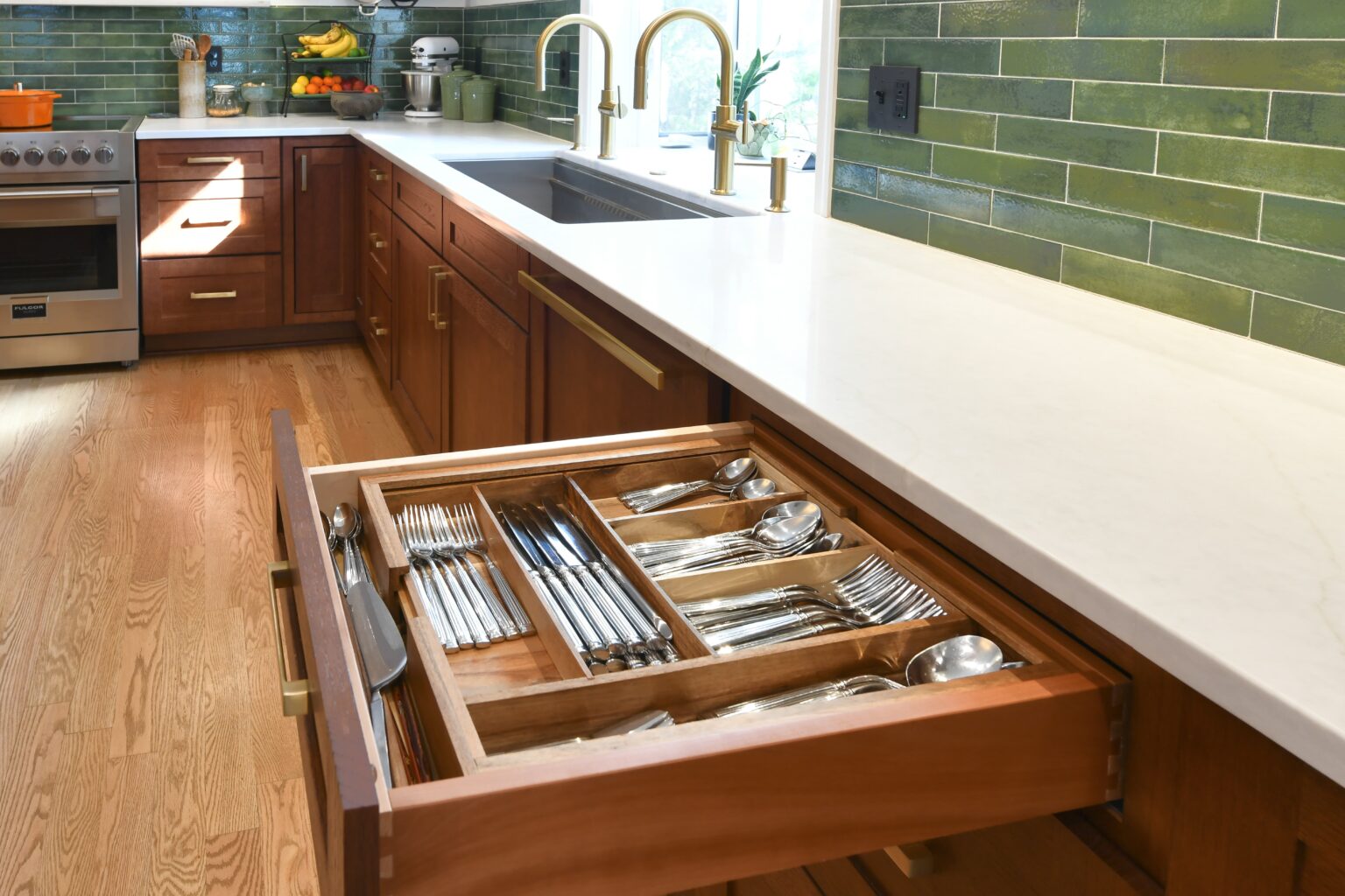 Open kitchen drawer with organized silverware in a wooden tray, set in a modern kitchen with green tile backsplash and gold faucets.