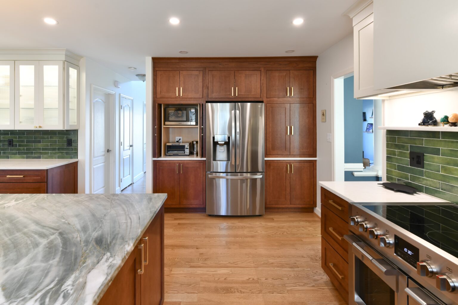 Spacious modern kitchen with stainless steel appliances, wood cabinets, green tile backsplash, marble island, and hardwood floors under bright recessed lighting.