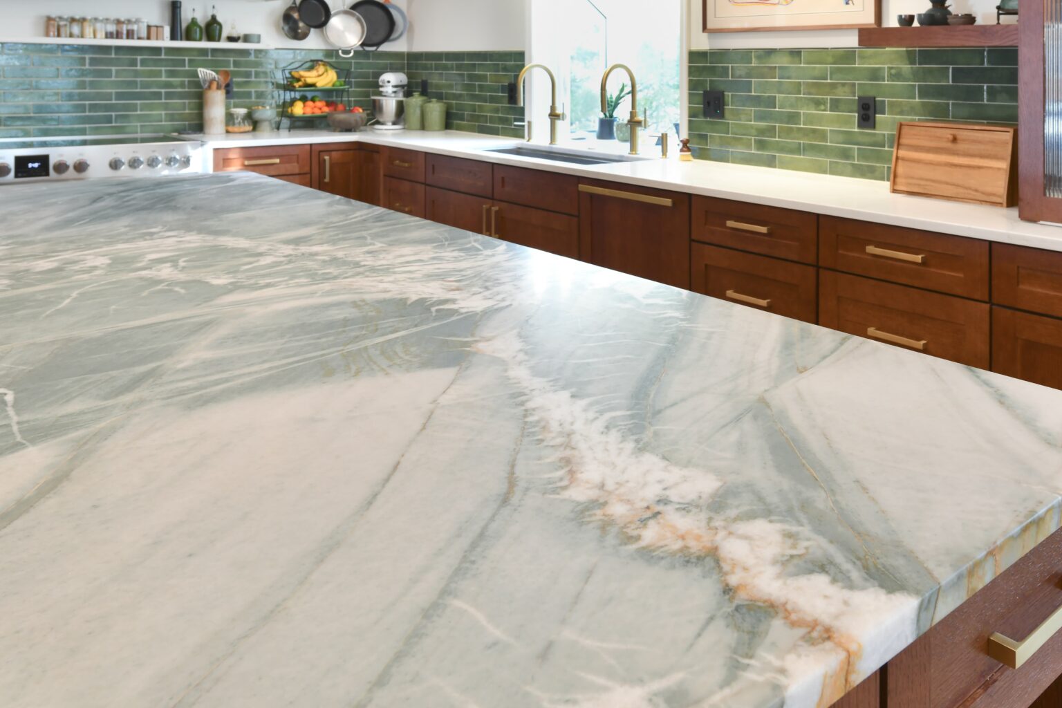 Close-up of a marble kitchen island with natural veining; green tile backsplash, wood cabinets, and brass fixtures in the background.