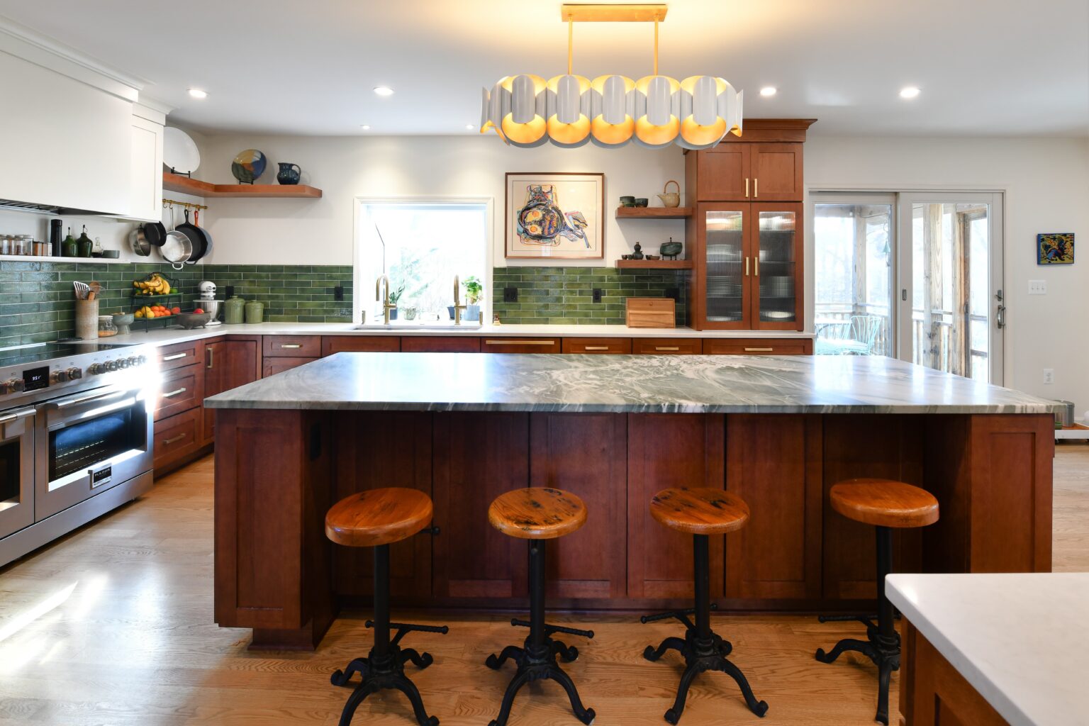 Spacious kitchen with a large island, wood cabinets, green tile backsplash, stainless steel appliances, and three wooden barstools.
