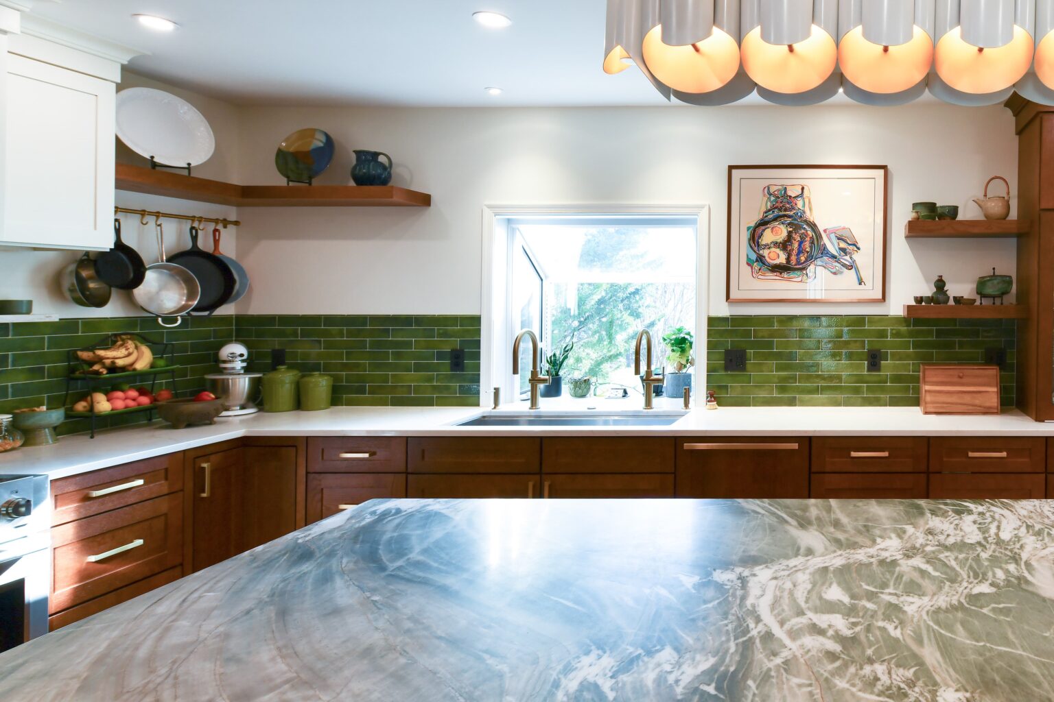 Modern kitchen with green tile backsplash, wooden cabinets, marble island, shelves with dishes, and a large window above the sink.
