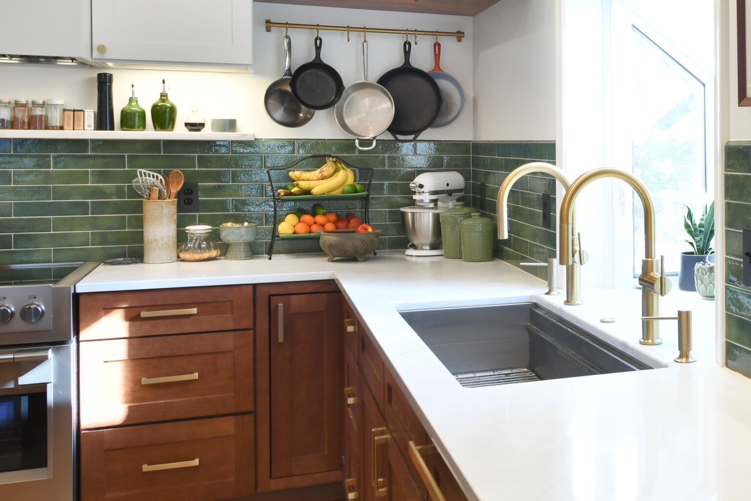 Modern kitchen with green tile backsplash, wooden cabinets, gold fixtures, hanging pans, countertop appliances, and a fruit bowl.